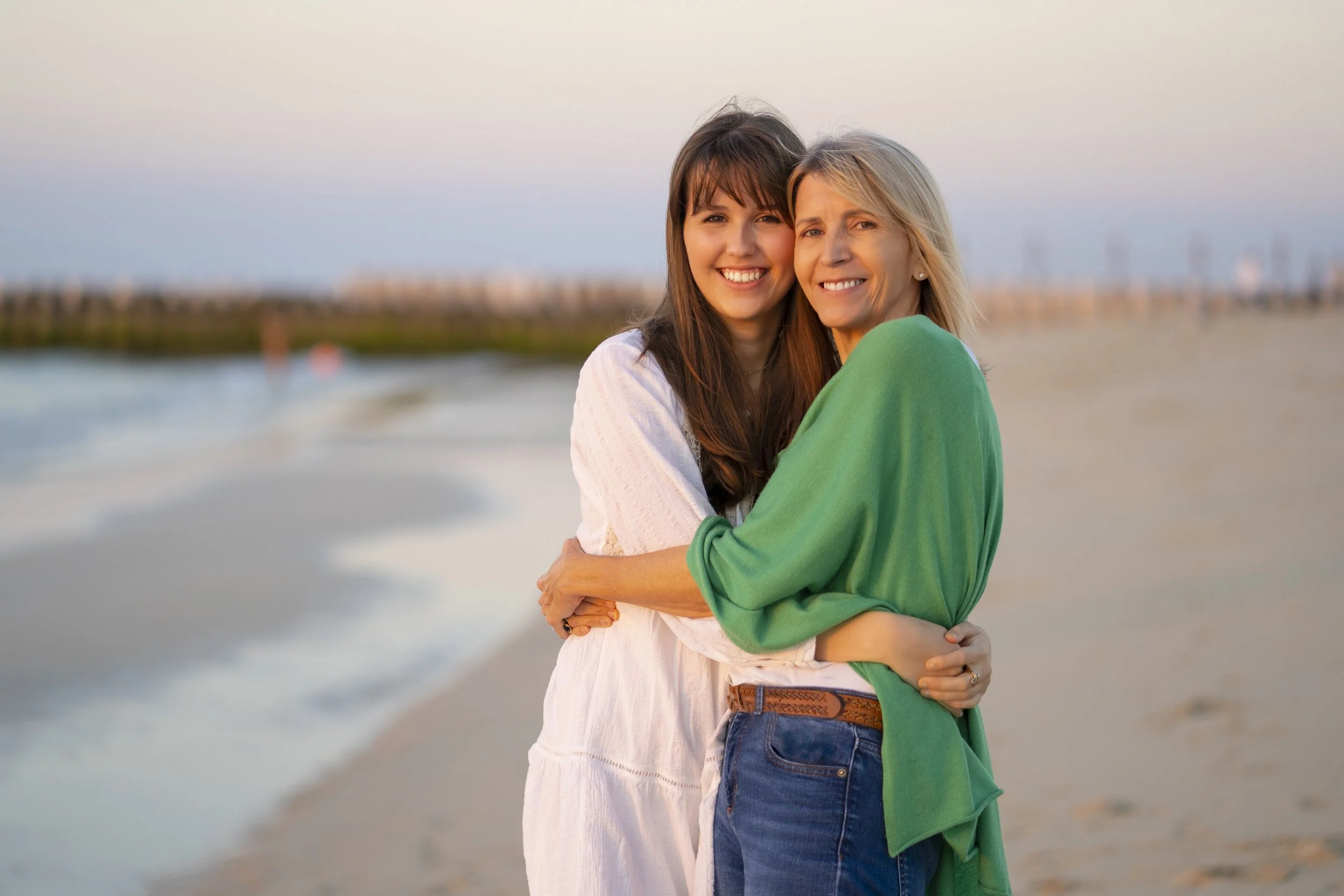 A young woman and an older woman hugging and smiling on a beach during sunset.