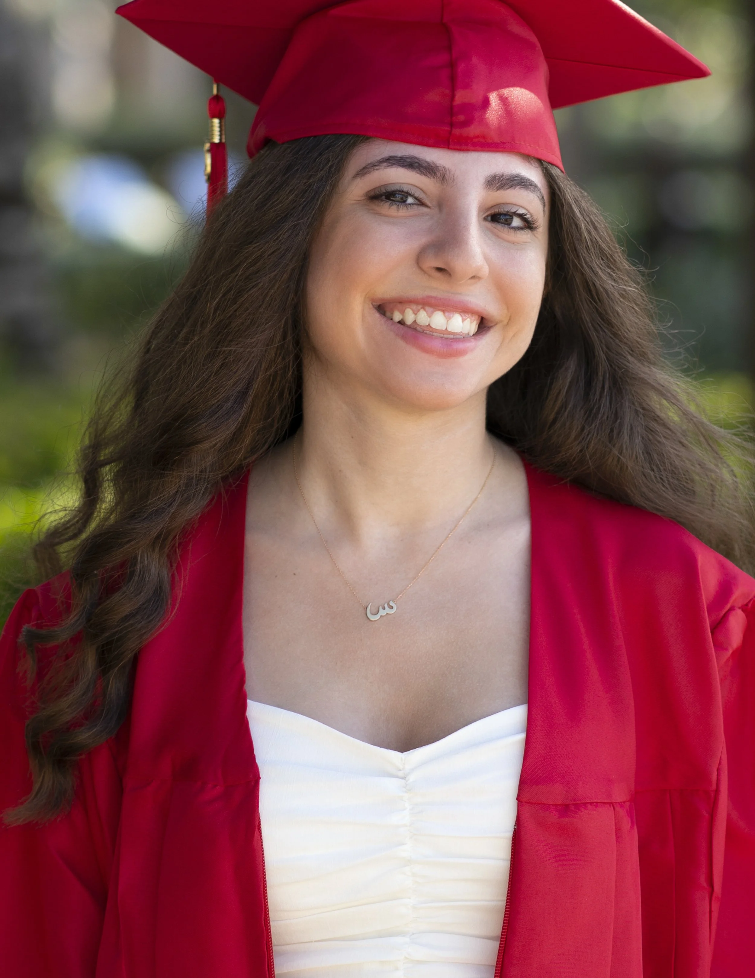 A young woman in a red graduation cap and gown, smiling outdoors with a blurred background of greenery.