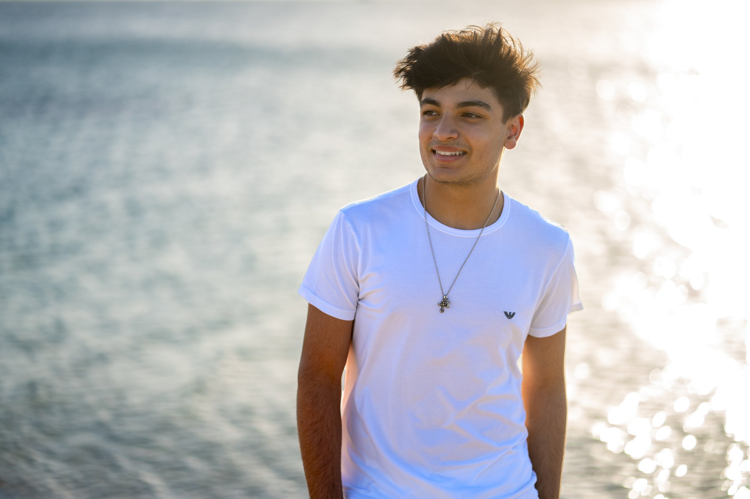 A young man with dark, tousled hair smiling while standing near water during sunset, wearing a white t-shirt and a silver necklace.