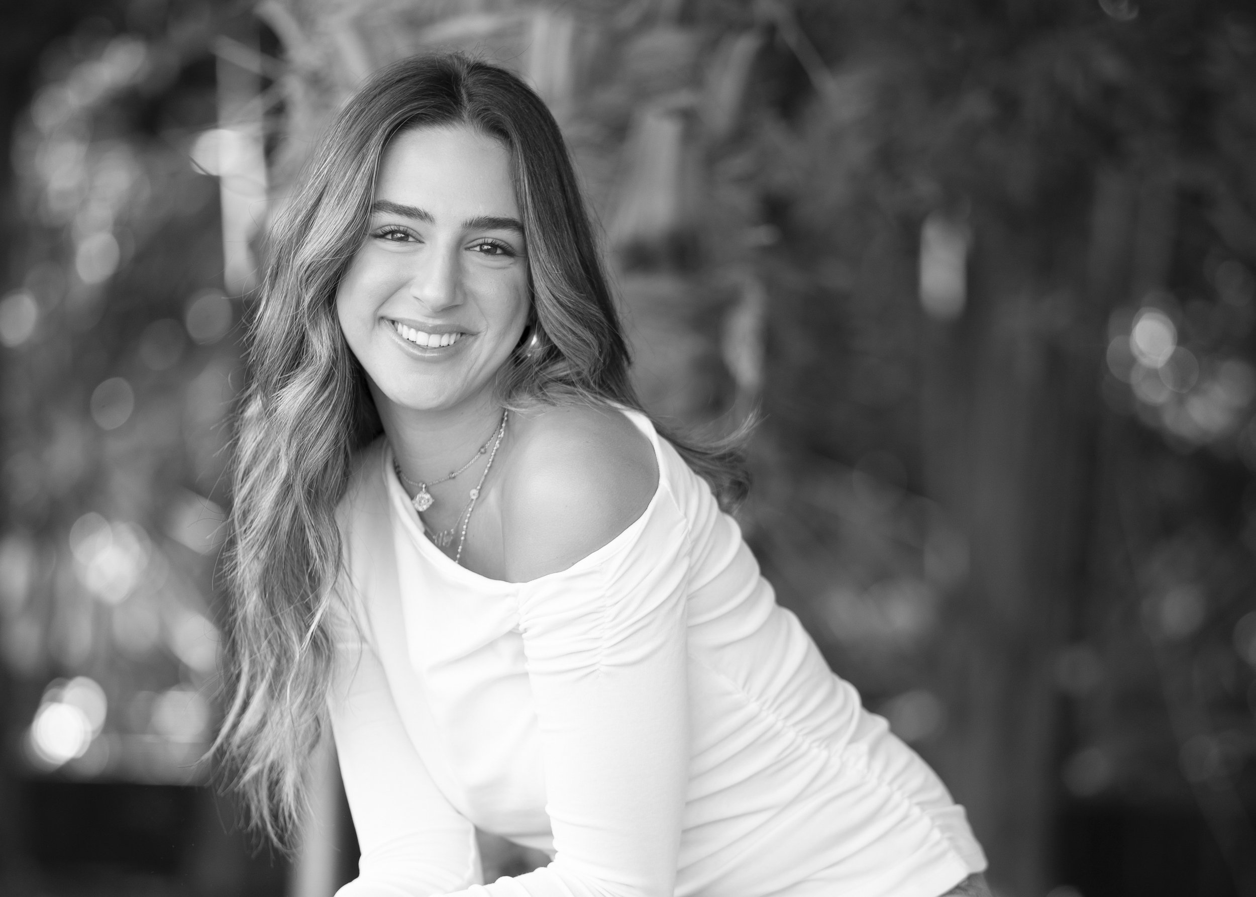 A smiling young woman with wavy hair, wearing a light-colored top and layered necklaces, outdoors with bokeh background.