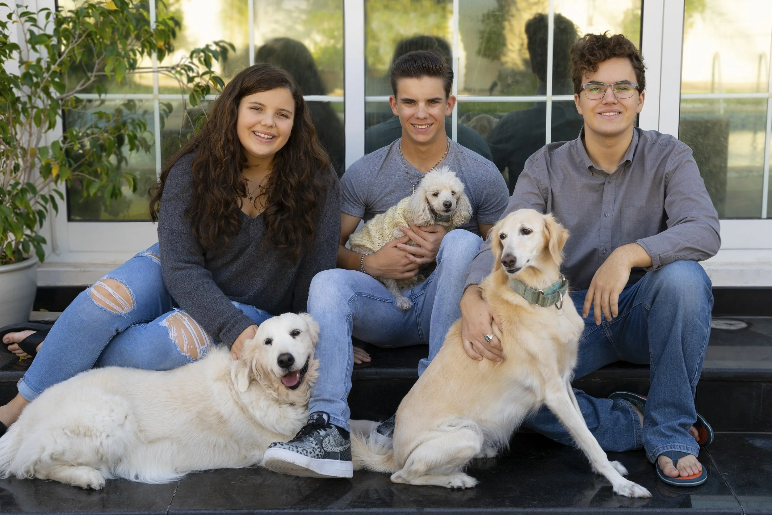 Three young people sitting on a black platform with two dogs, outside near a glass building, smiling for a group photo.
