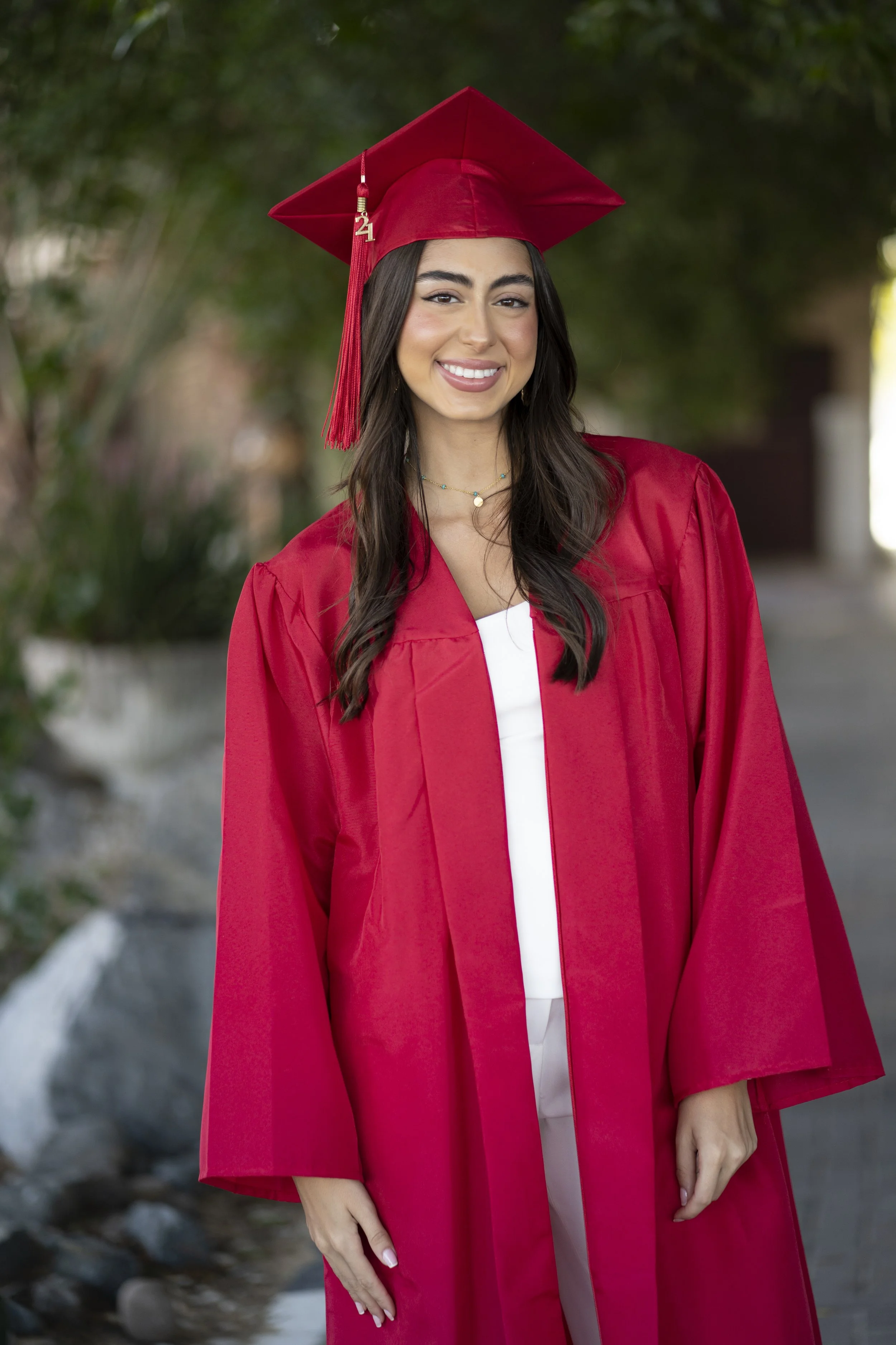 A young woman in a red graduation gown and mortarboard cap smiling outdoors.