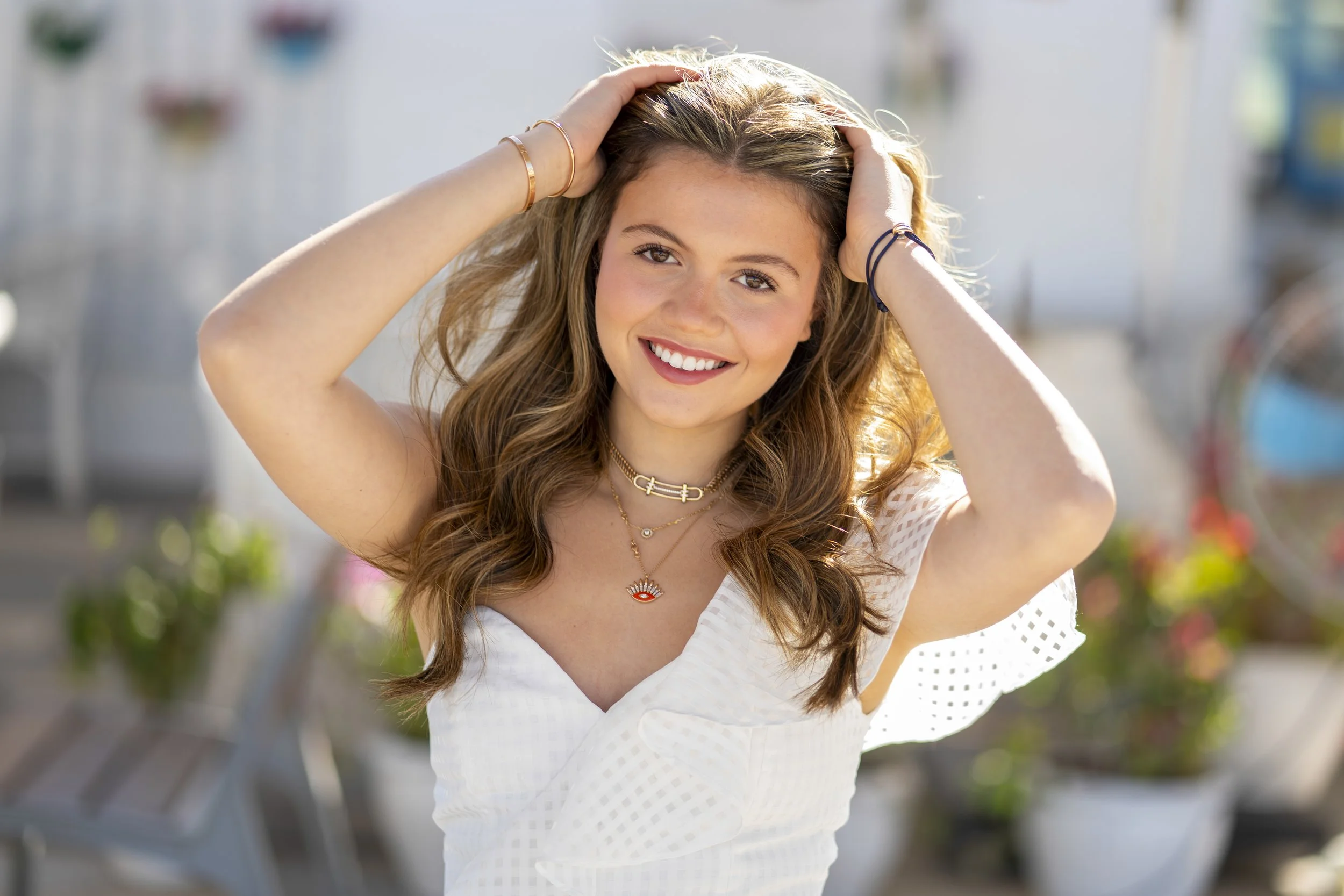 A young woman with long, wavy brown hair, smiling, wearing a white dress with polka dot sleeves and multiple necklaces, standing outdoors in bright sunlight.
