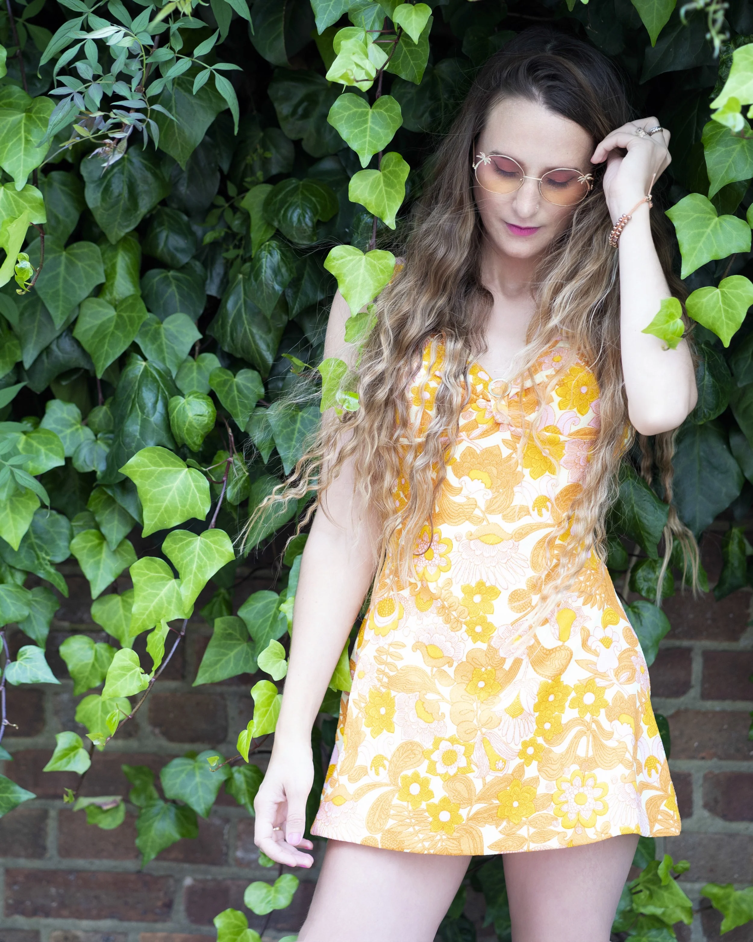 A woman with long wavy hair wearing pink sunglasses and a floral yellow and orange dress, standing against a lush green ivy-covered brick wall.