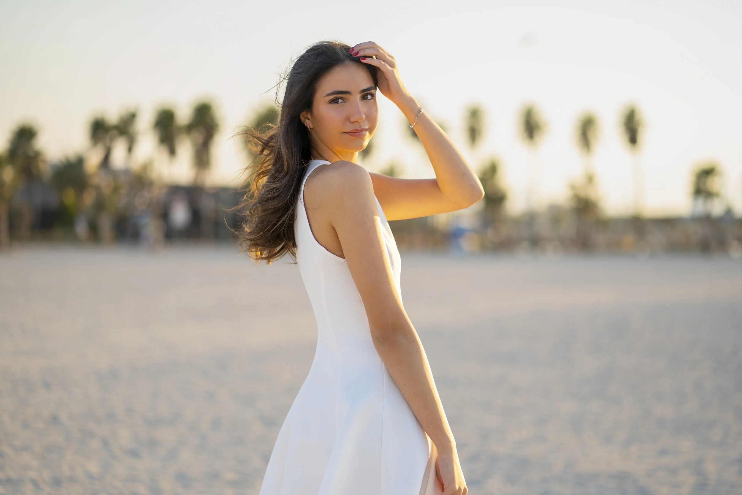 A young woman in a white dress standing on the beach at sunset, with palm trees in the background.