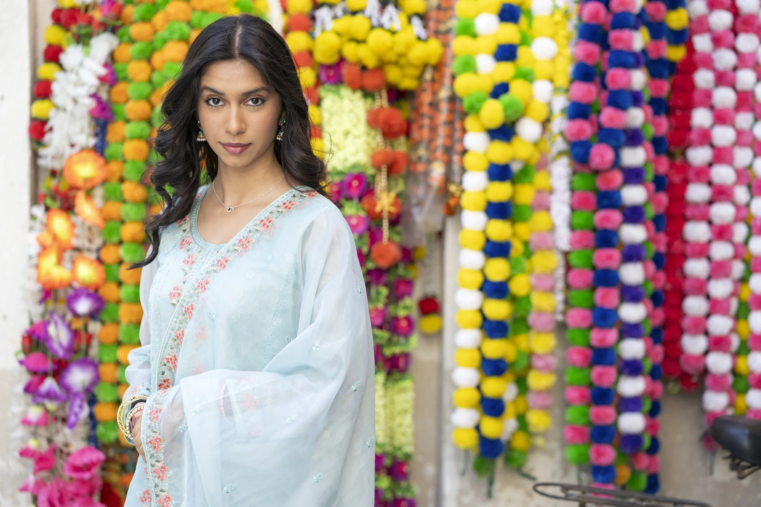 A woman with long dark hair and wearing a light blue embroidered dress stands in front of a colorful display of hanging flower garlands.