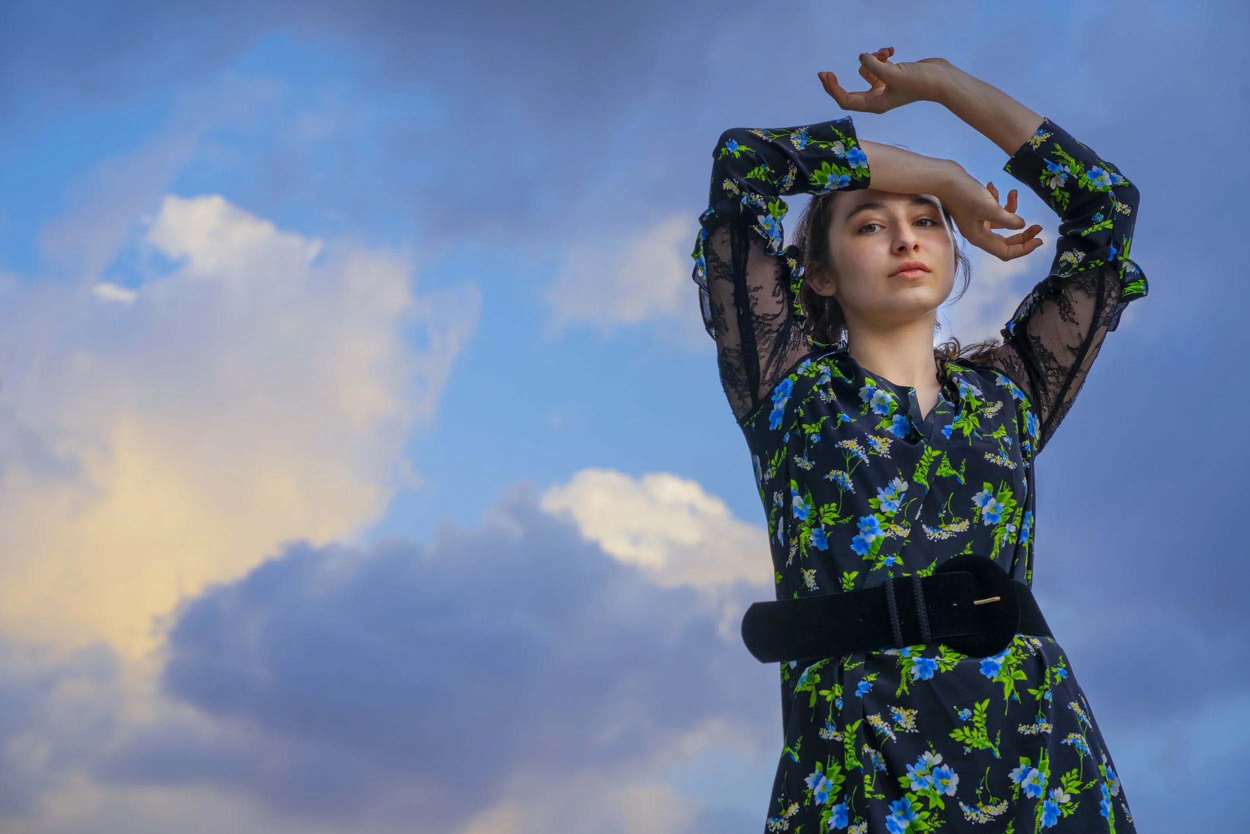 A woman with styled hair and neutral expression, wearing a floral dress with lace sleeves and a black belt, stands against a partly cloudy sky.