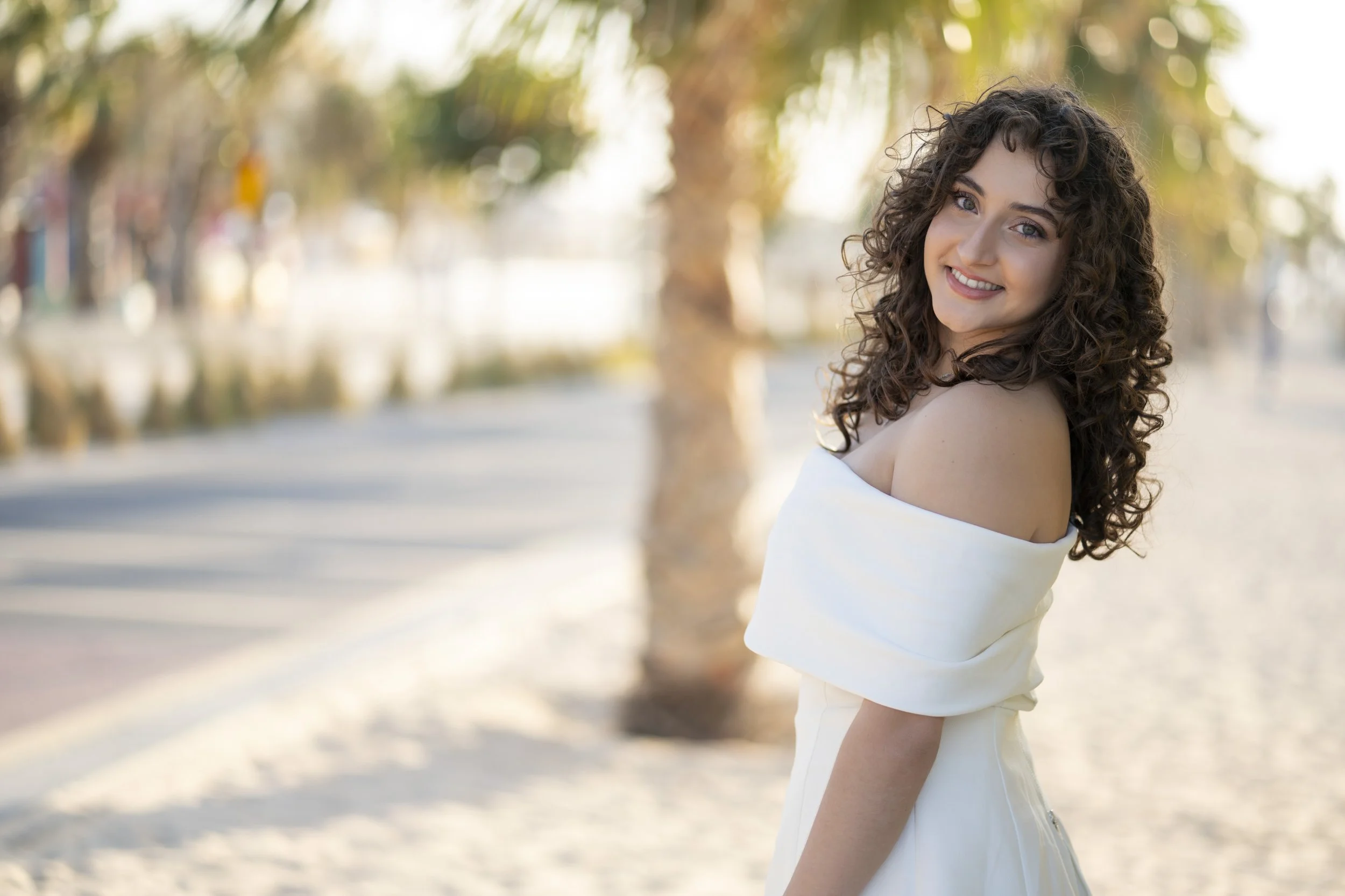 A young woman with long, curly hair is smiling at the camera while standing outdoors on a sandy path with trees in the background during the daytime. She is wearing an off-the-shoulder white dress.