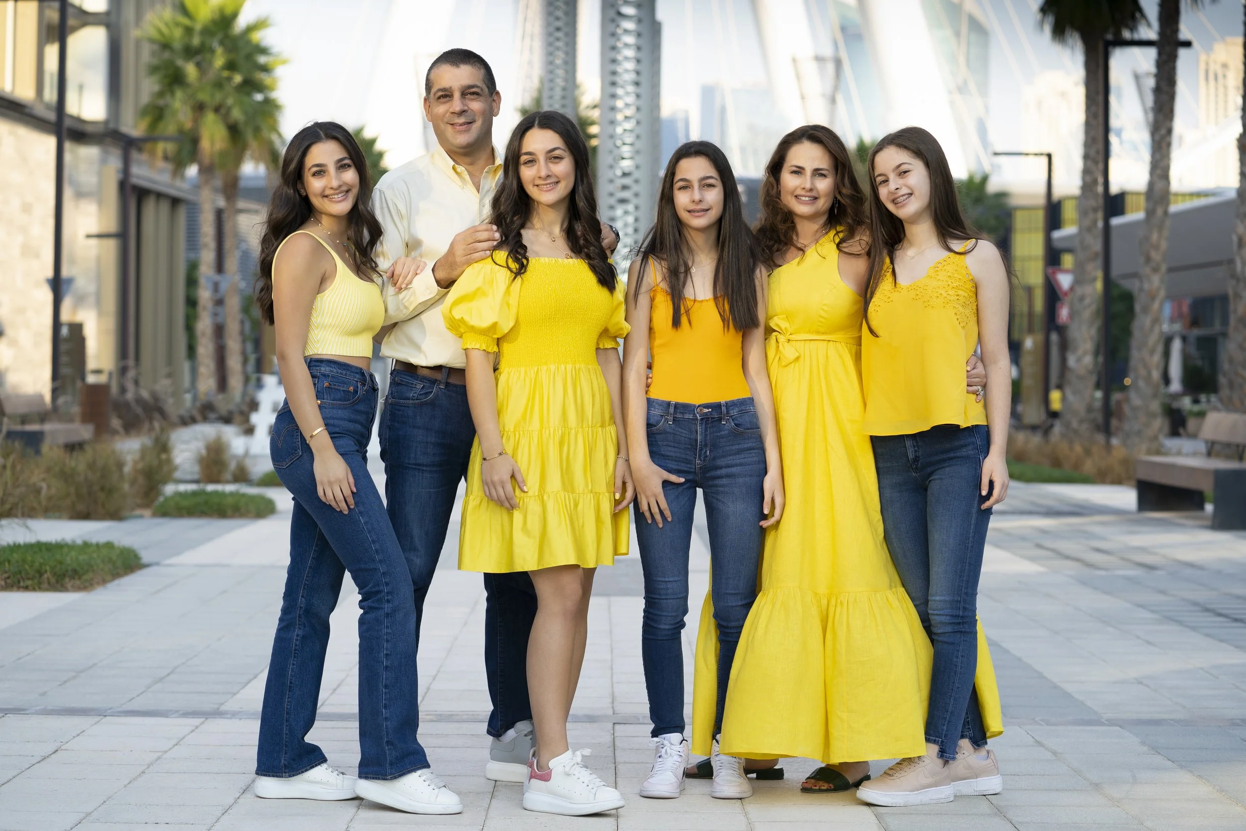 Family group of six people, three women, one man, and two girls, standing outdoors in a city setting, wearing yellow and casual clothing, smiling and posing for the photo.
