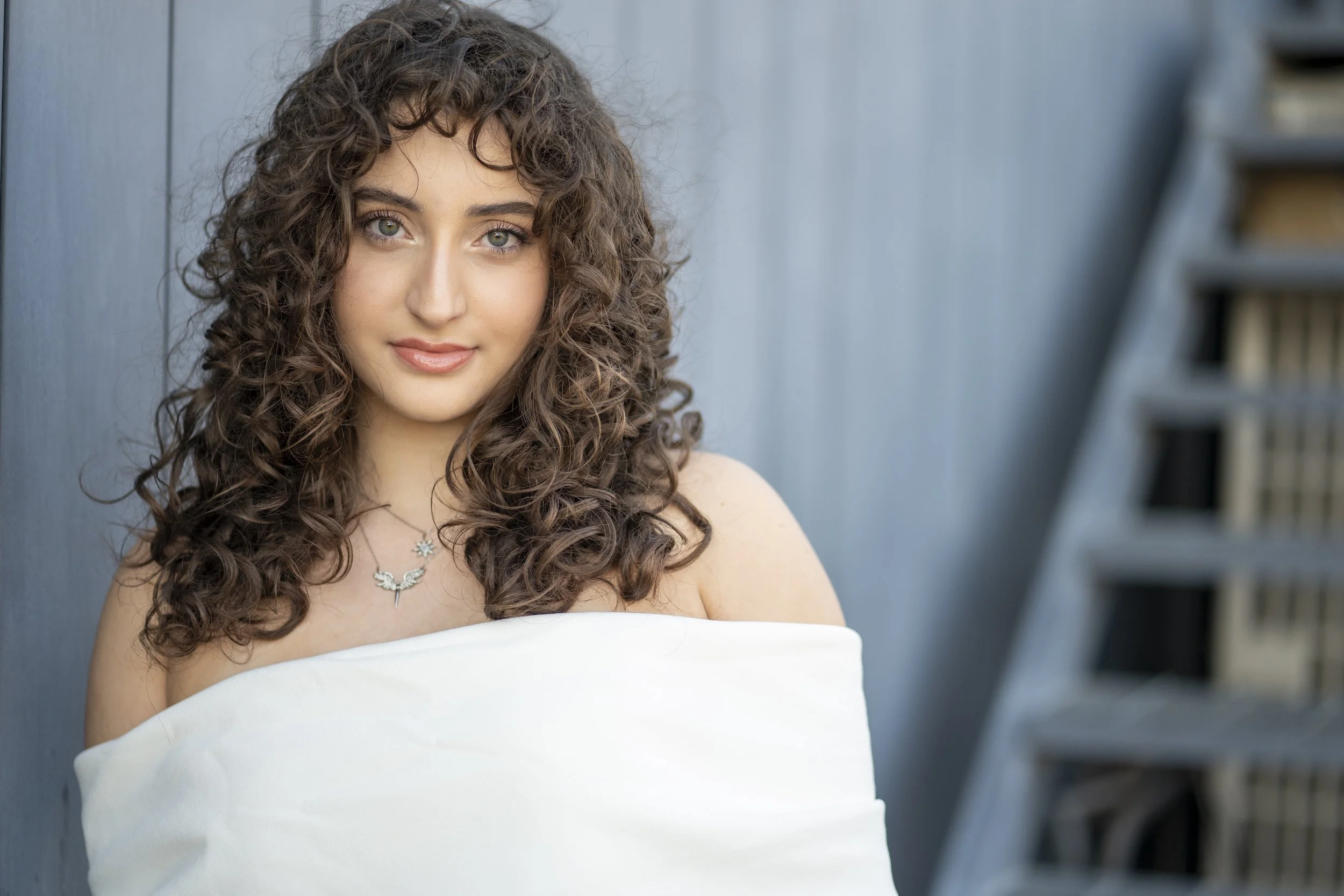 A young woman with curly brown hair and green eyes, smiling softly, wearing a necklace and a white off-shoulder top, standing against a blue wall with a staircase in the background.