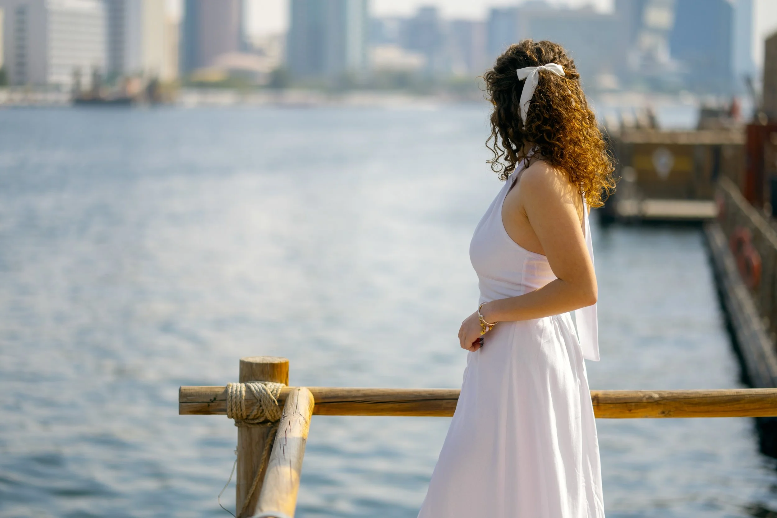A woman with curly brown hair tied with a white ribbon, wearing a white sleeveless dress, standing on a wooden dock by the water, with a city skyline in the background.