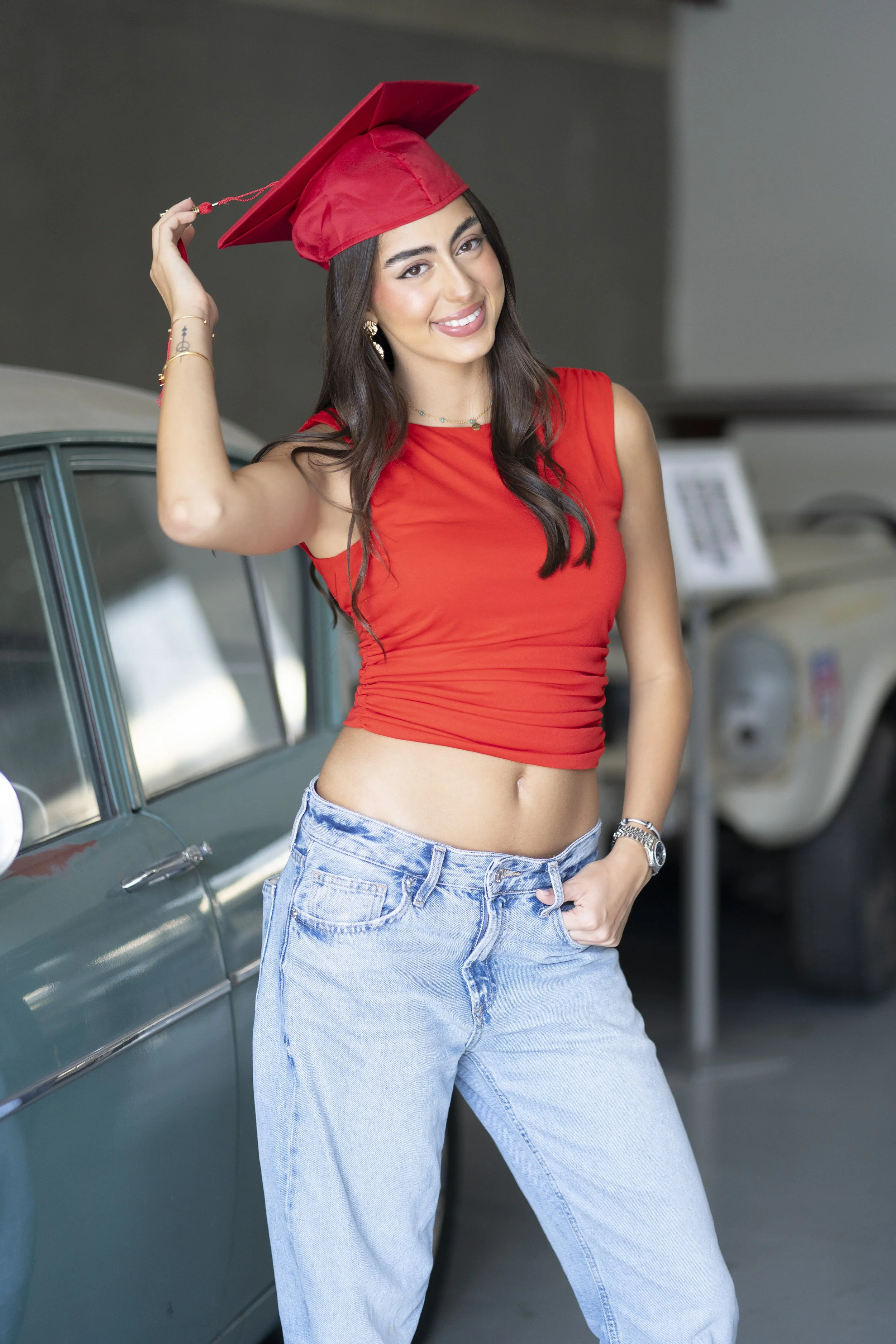 Young woman in red crop top and jeans posing with a red graduation cap in a garage, smiling.