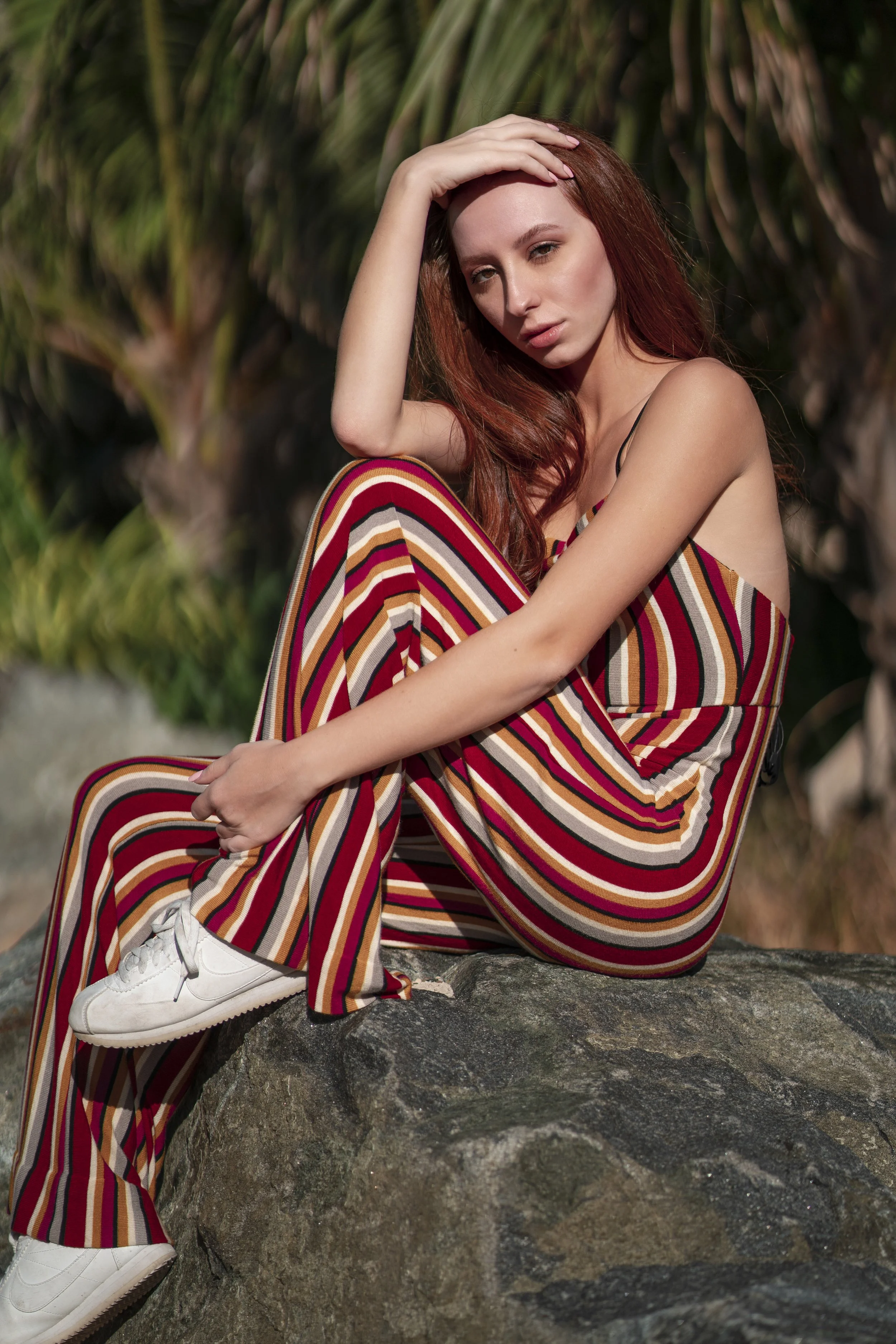 A woman with long red hair sitting on a rock outdoors, wearing a striped jumpsuit and white sneakers, with one hand on her head and the other on her knee.