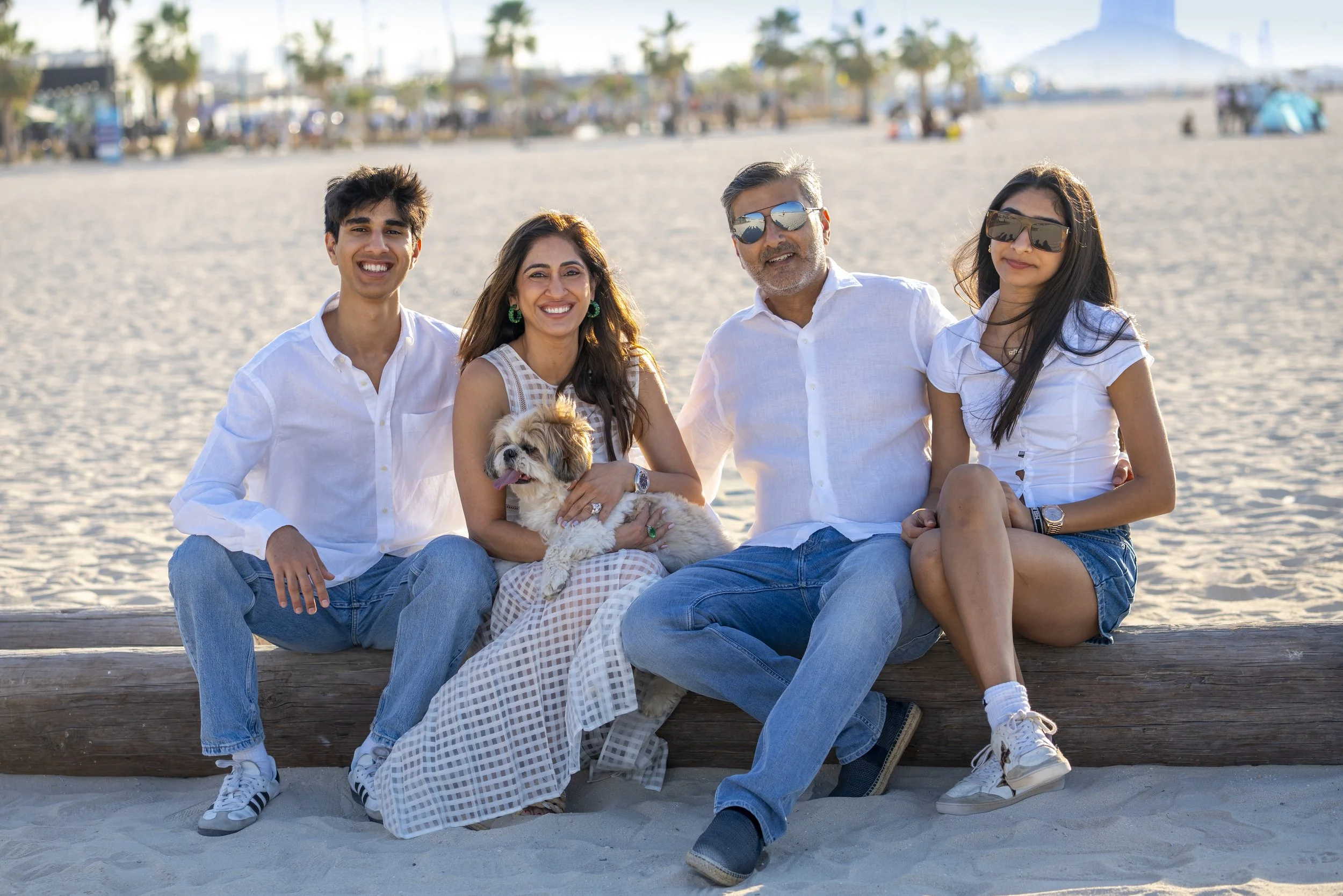 A family of five, including a dog, sitting on a wooden bench on a sandy beach with umbrellas, palm trees, and people in the background, enjoying a sunny day.