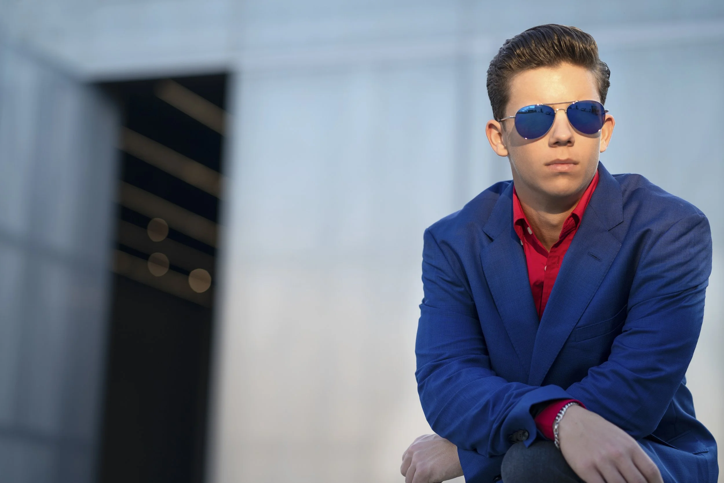 Young man with slicked-back hair wearing blue sunglasses, a blue blazer, and a red shirt, sitting outdoors with modern architecture in the background.