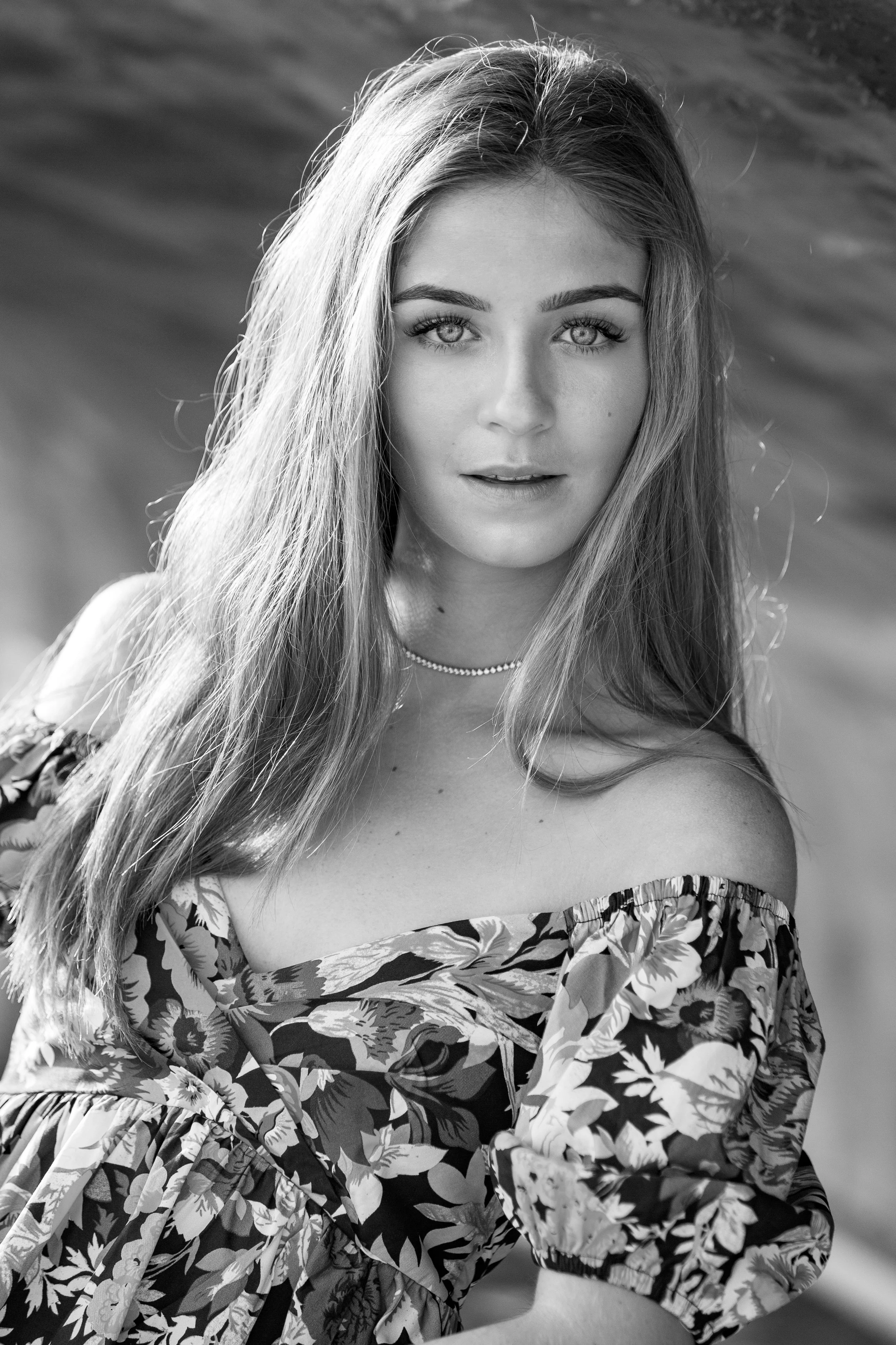 Black and white photo of a young woman with long hair, wearing a floral off-the-shoulder dress and a pearl necklace.