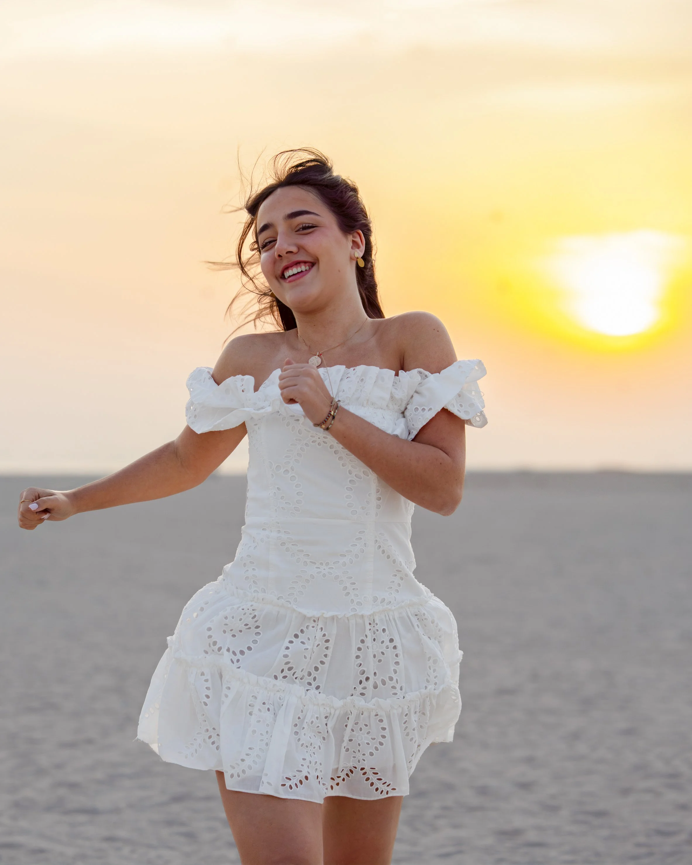 A woman is running on the beach during sunset, wearing a white off-shoulder dress and smiling.