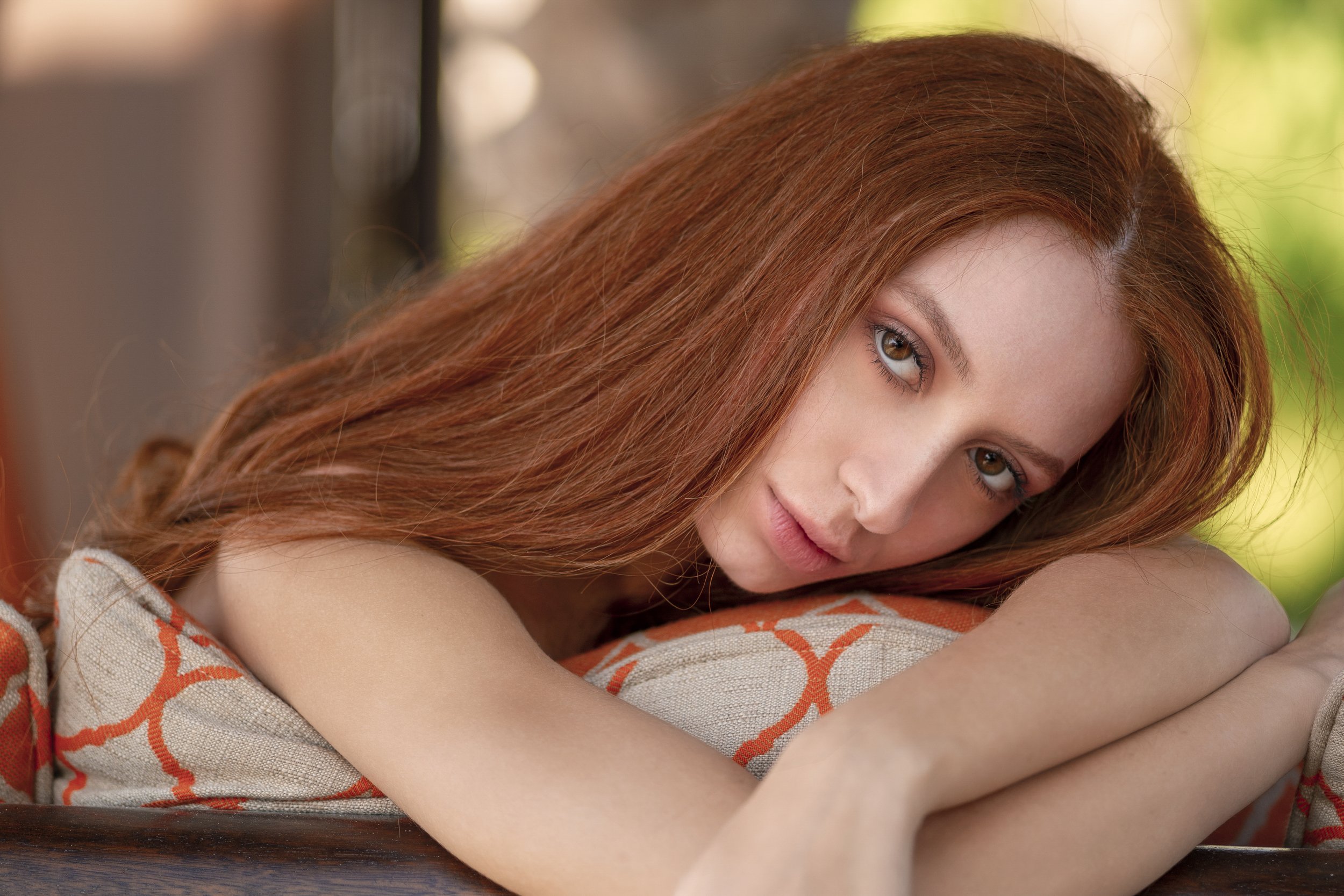 A young woman with long red hair and light skin resting her head on her arms, lying on a patterned cushion with orange and beige geometric designs, gazing softly at the camera.
