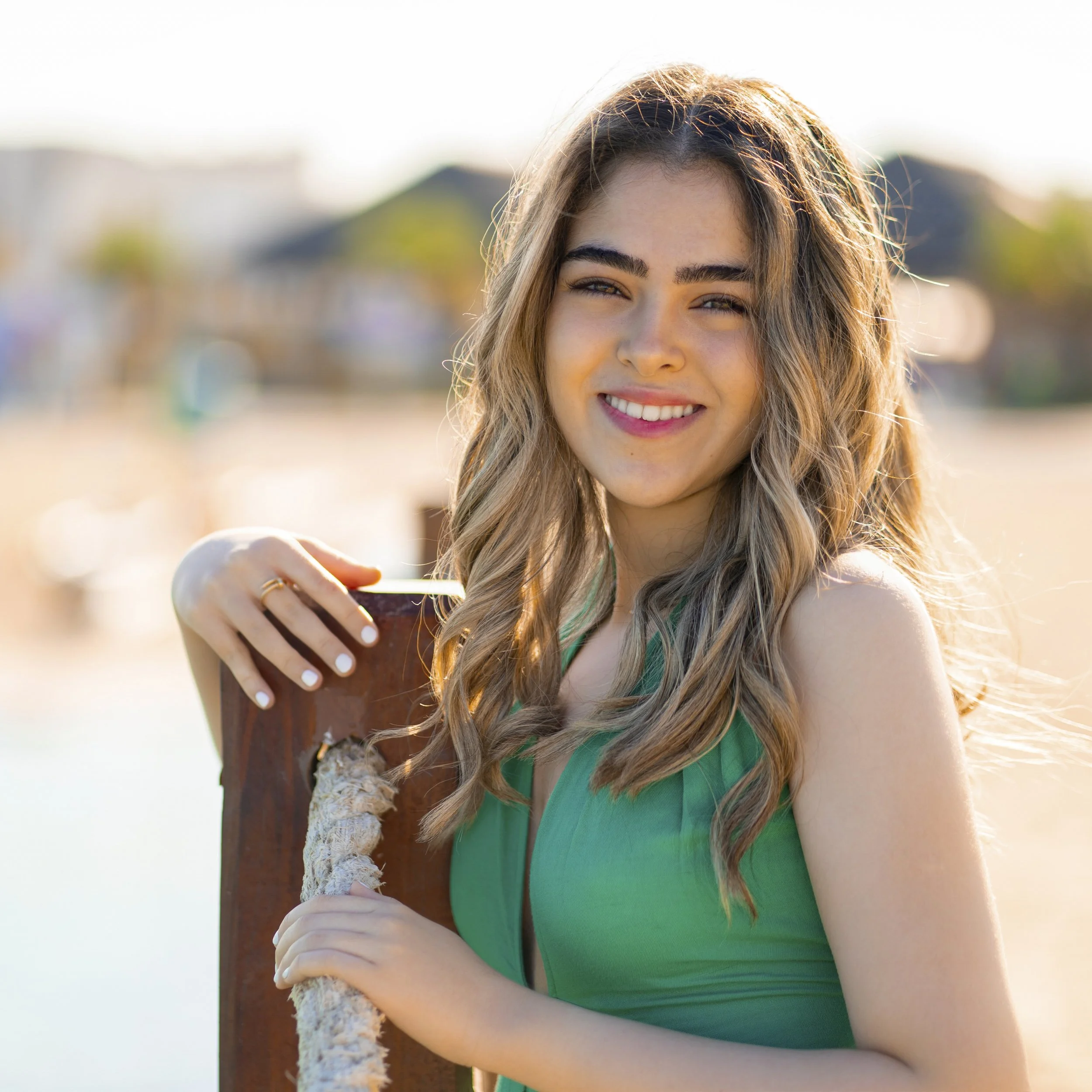 A young woman with long, wavy blonde hair smiling at the camera, standing outdoors on a beach near water with blurred buildings in the background, wearing a green sleeveless top.