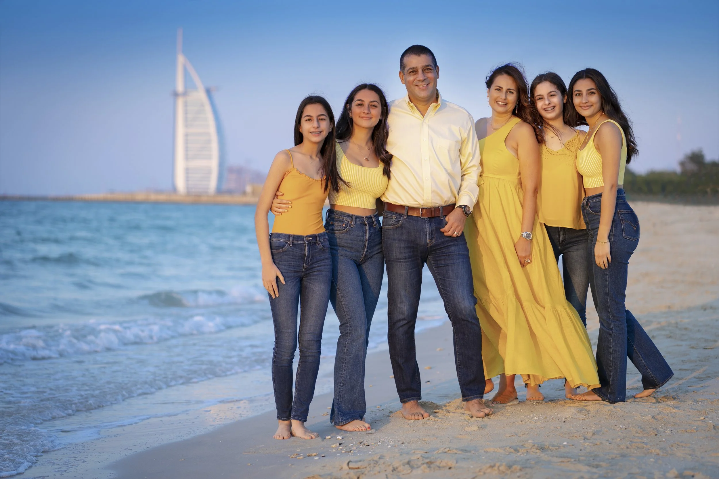 Family of six standing on the beach in front of the ocean with the Burj Al Arab hotel in Dubai in the background. They are smiling, wearing yellow tops and blue jeans, with some barefoot on the sand.