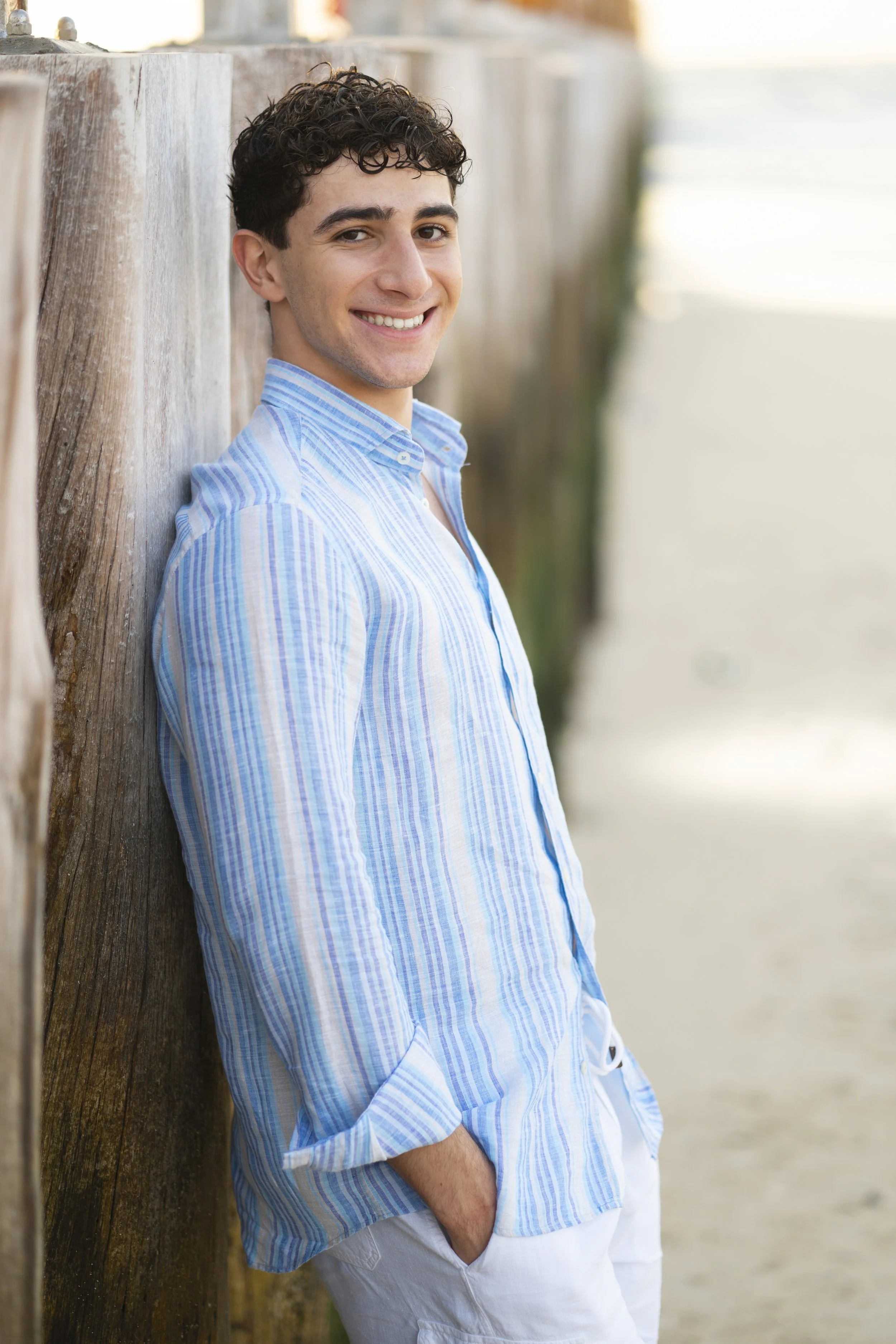 A young man with curly dark hair, smiling, leaning against a wooden structure on a beach, wearing a blue striped button-up shirt with rolled-up sleeves and white shorts.
