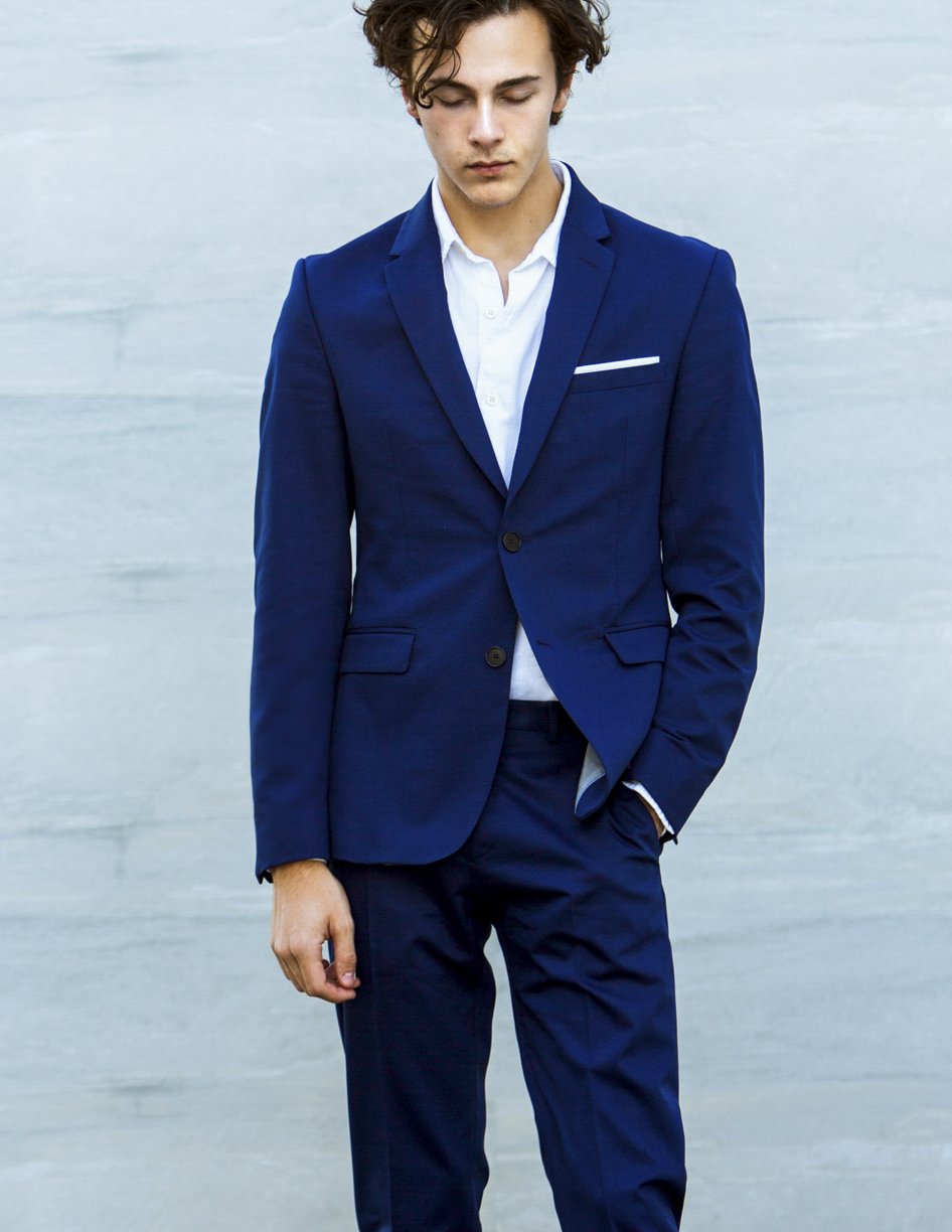 Young man in a navy blue suit with a white shirt, standing with his eyes closed against a light background.