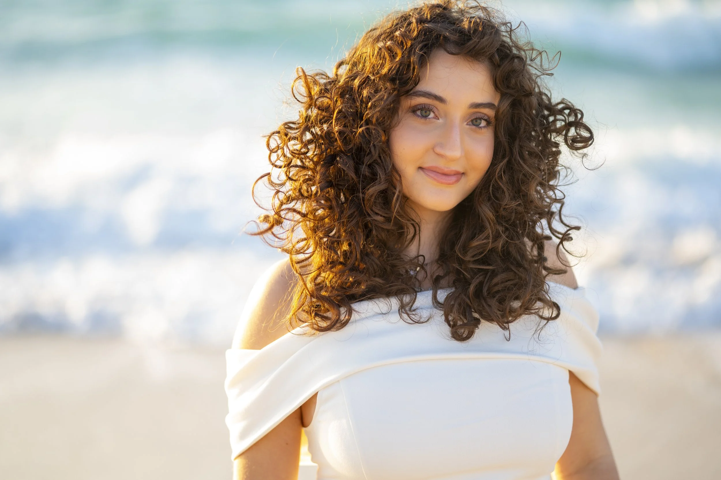 A young woman with curly brown hair and light skin stands on a beach, smiling gently at the camera with the ocean and waves in the background.