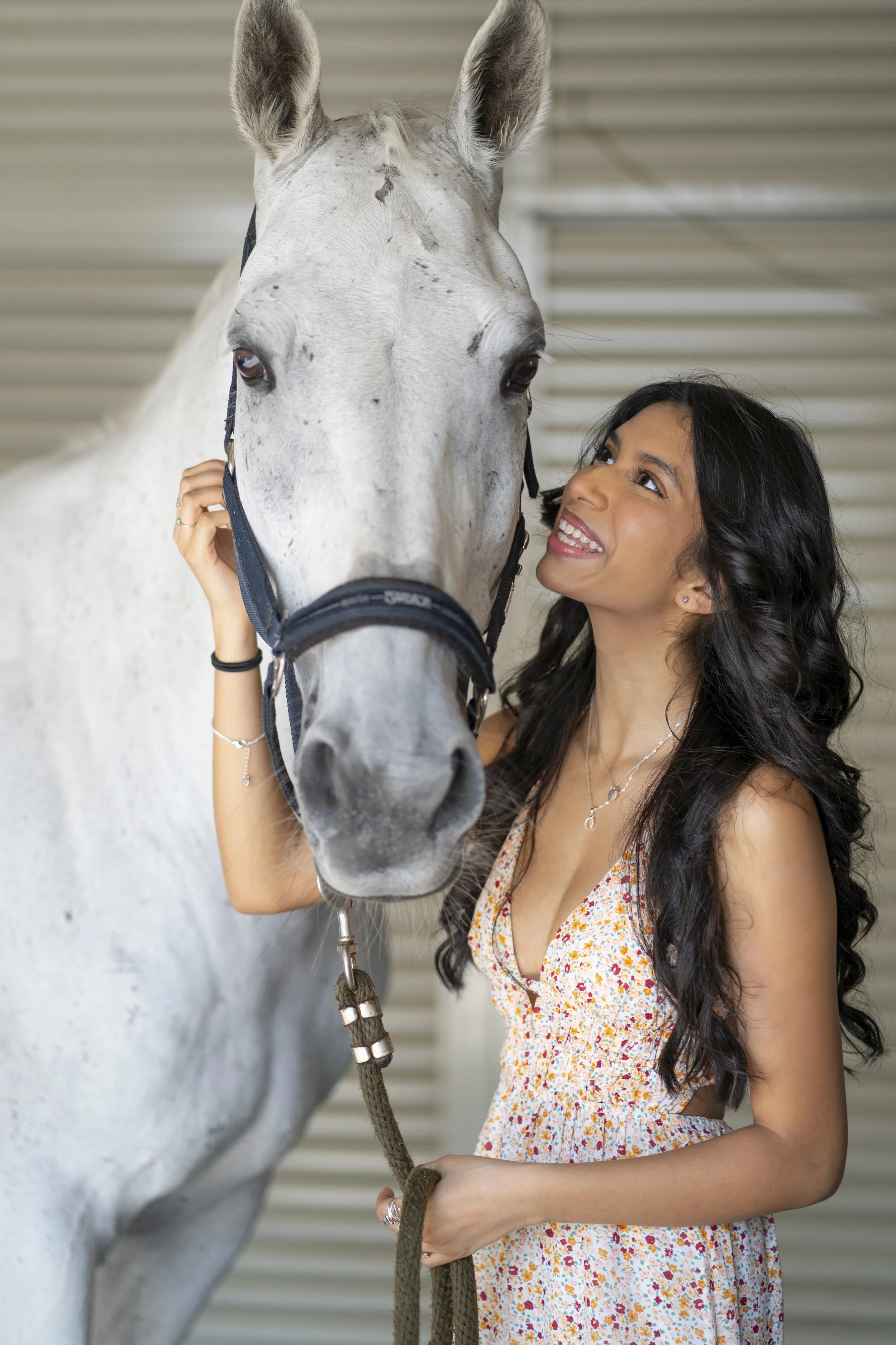 A woman with long dark curly hair smiling and looking at a white horse with gray spots, in a barn or stable.