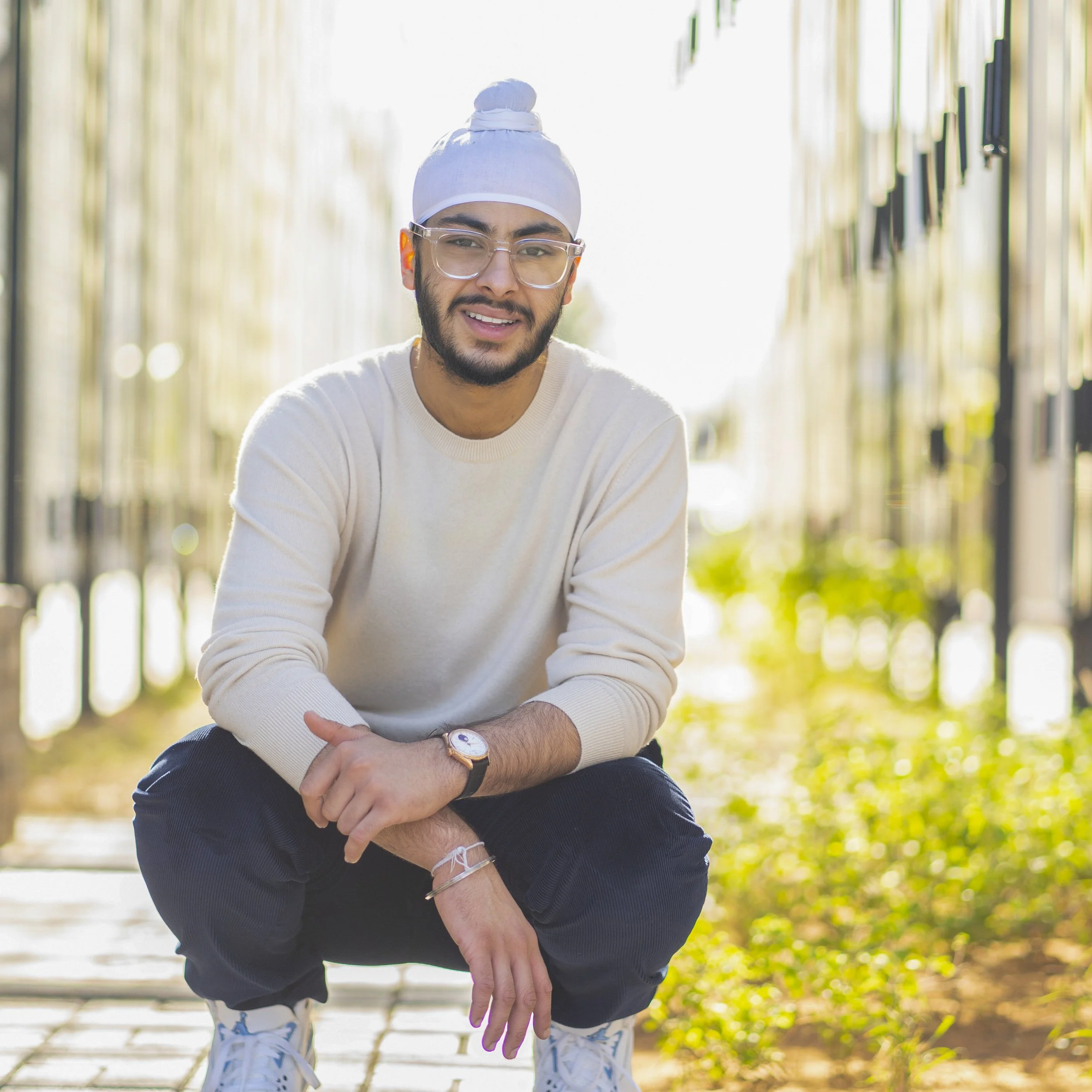 A young man with glasses and a white head wrap squatting outdoors on a sunny day, smiling at the camera.