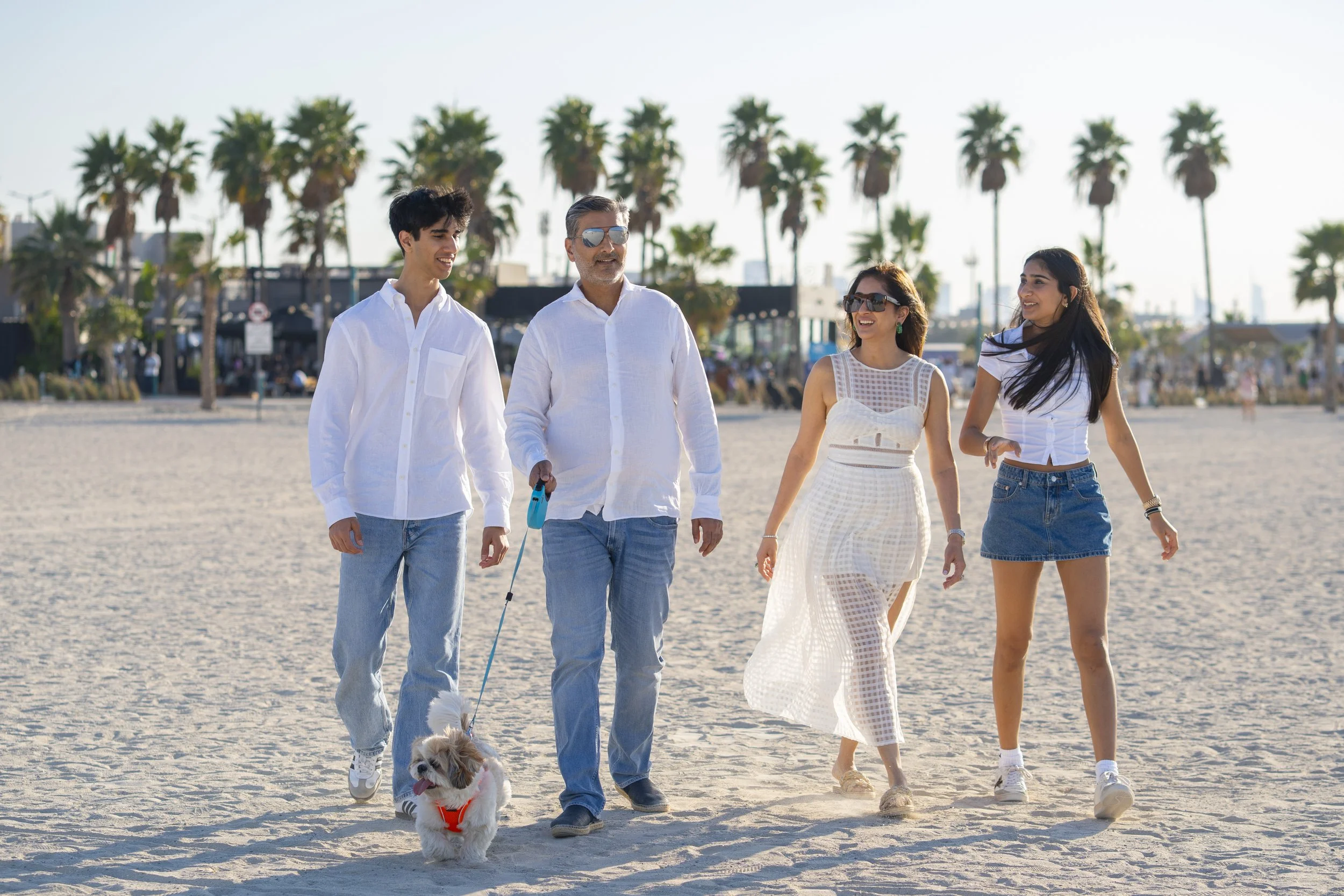 Four people walking on the beach, two men and two women, with palm trees and beachside structures in the background, a man with sunglasses is holding a small dog on a leash.