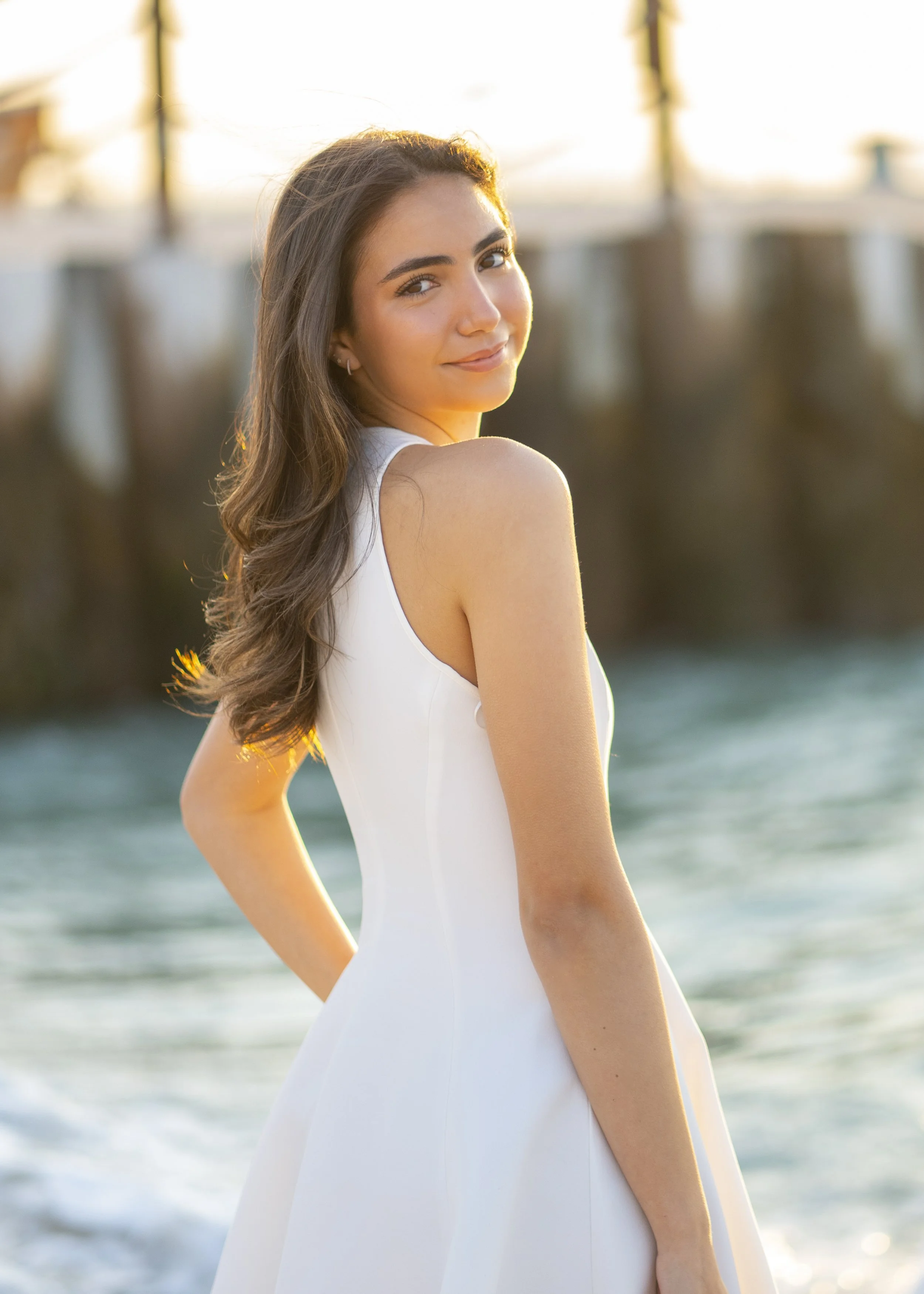 A young woman in a white dress standing by the water with a wooden pier in the background during sunset, smiling at the camera.