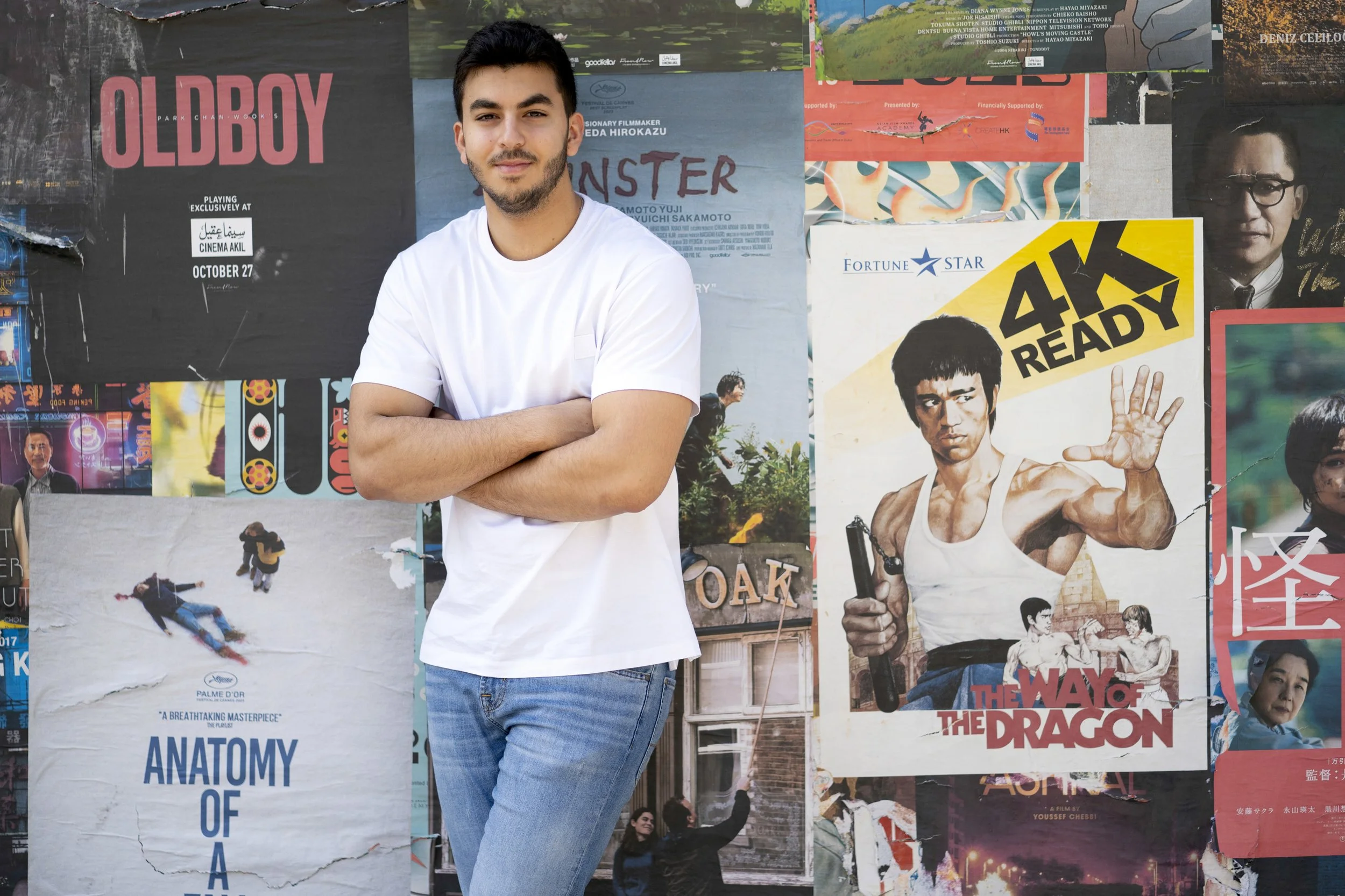 A young man with dark hair and a beard standing with crossed arms in front of a colorful wall of posters, including a prominent poster of Bruce Lee from the movie 'The Way of the Dragon' and another poster for the film 'Anatomy of a Murder.'