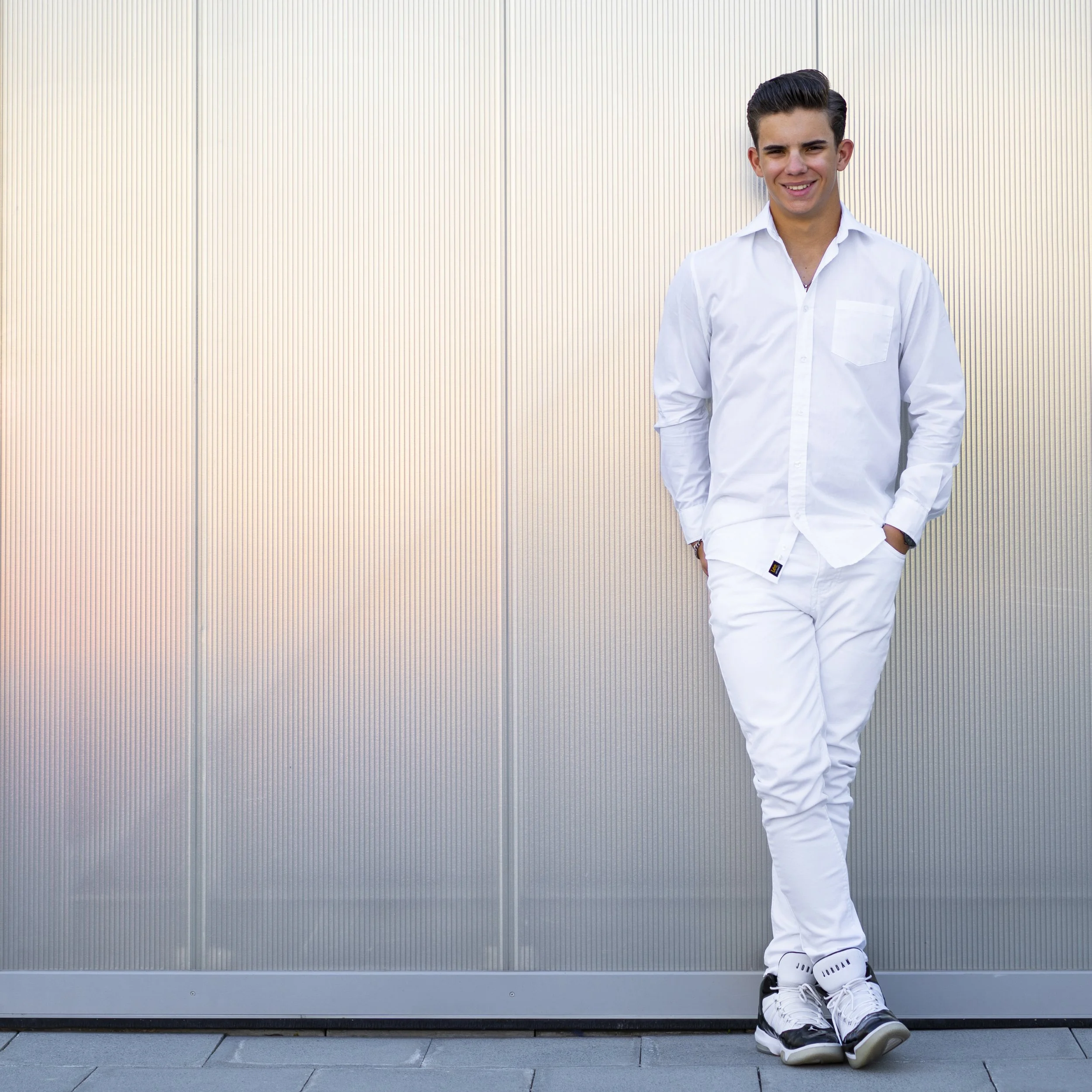 Young man in white clothing, standing outdoors against a metallic wall, smiling with hands in pockets, wearing sneakers.