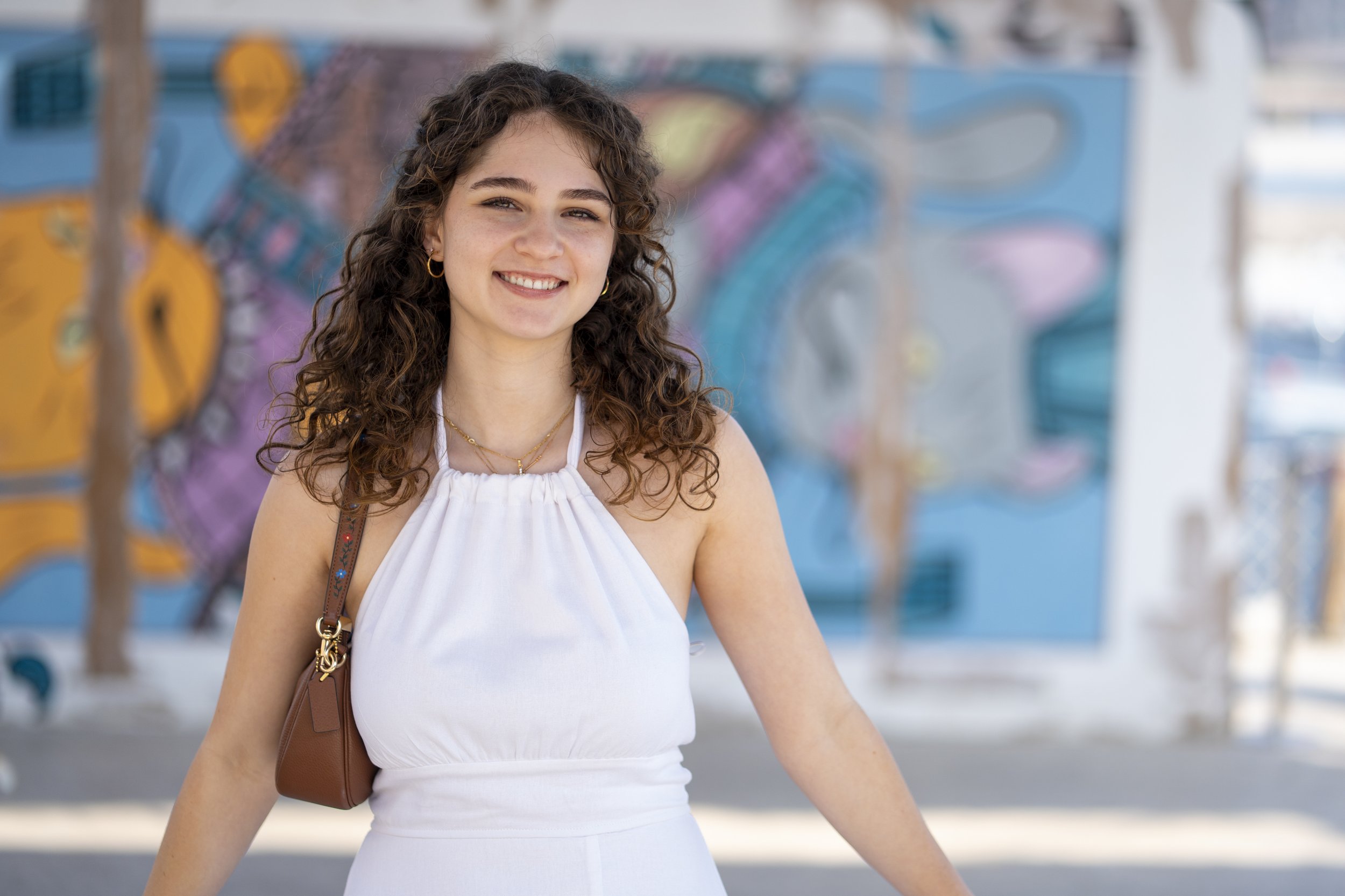 A young woman with curly brown hair smiling outdoors with a colorful graffiti wall in the background.