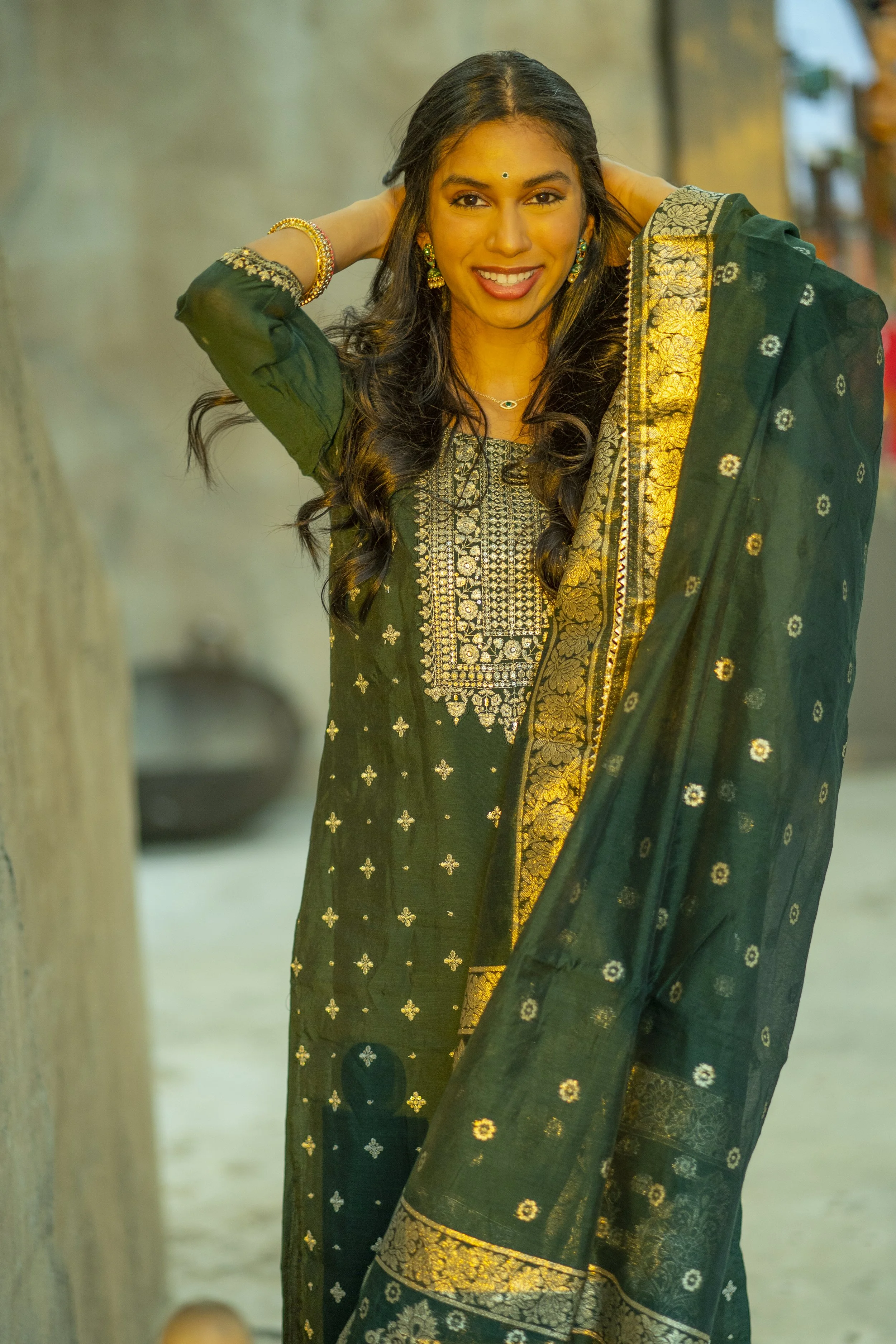 A woman dressed in traditional Indian attire, smiling and adjusting her hair, standing outdoors.