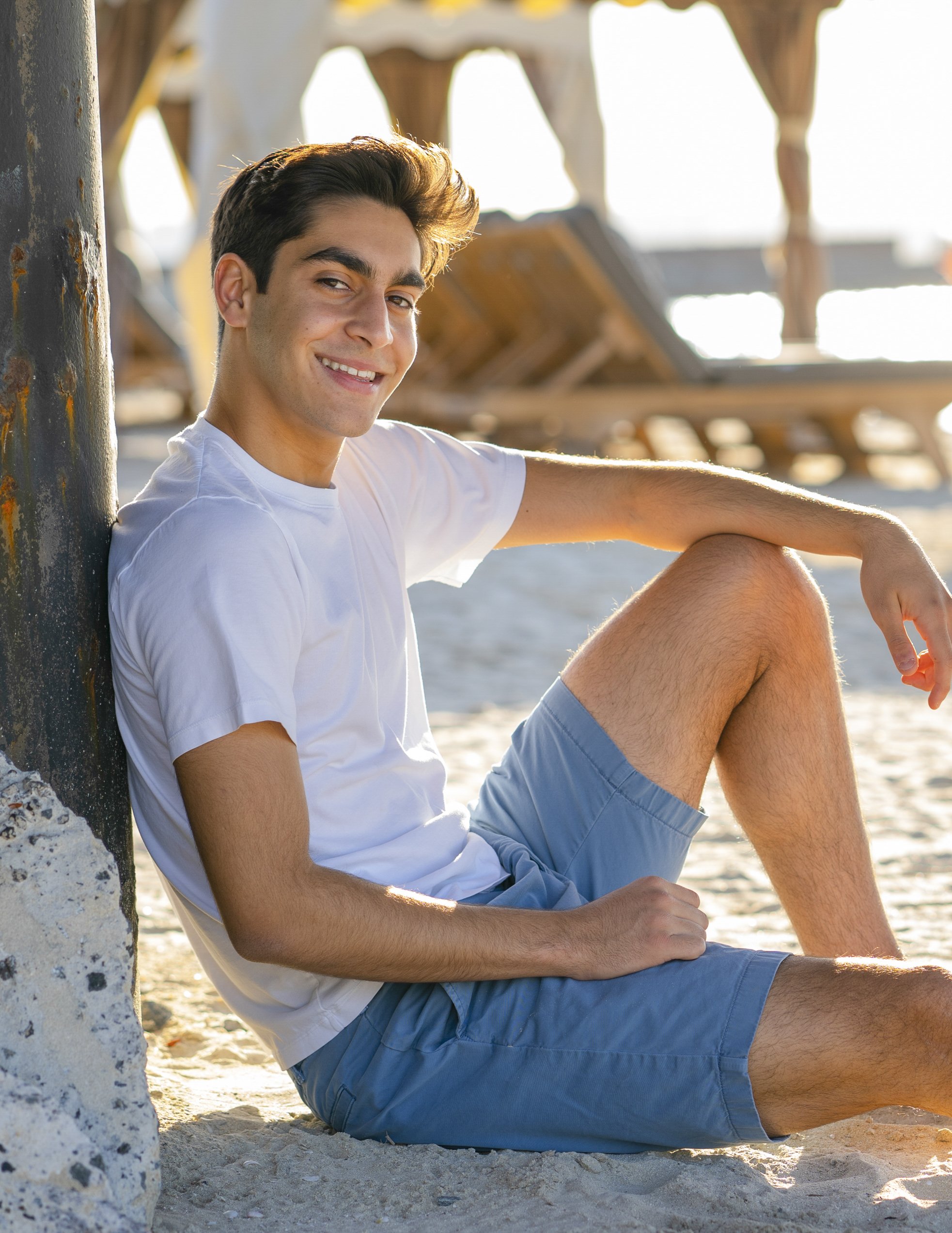 A young man sitting on the beach next to a wooden pier, smiling at the camera, wearing a white t-shirt and blue shorts.