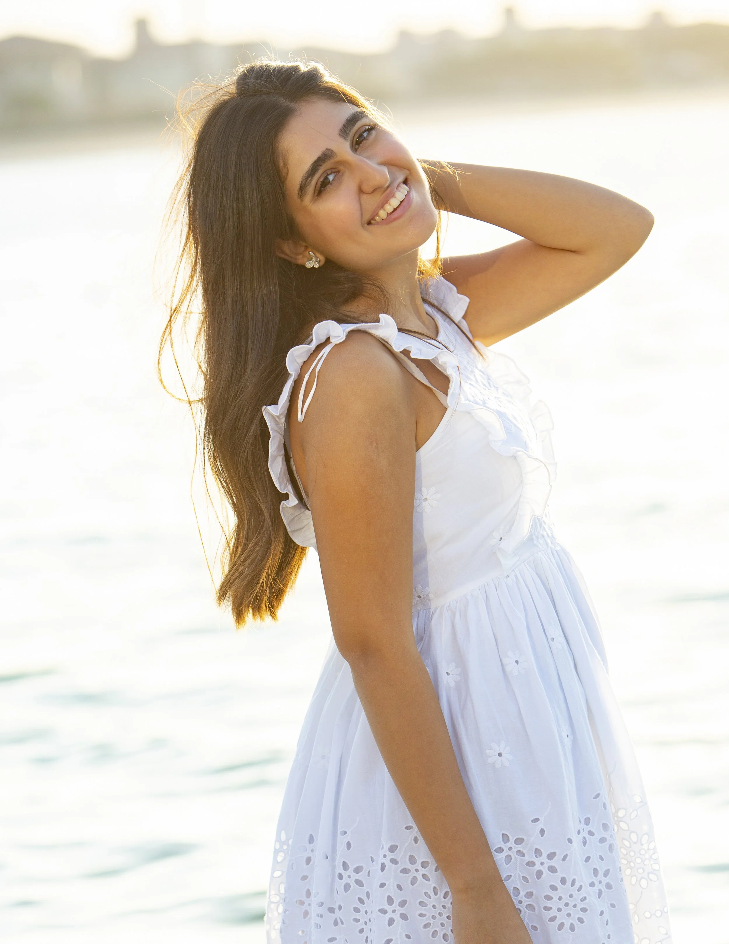 A young woman smiling at the camera at the beach during sunset, wearing a white dress.