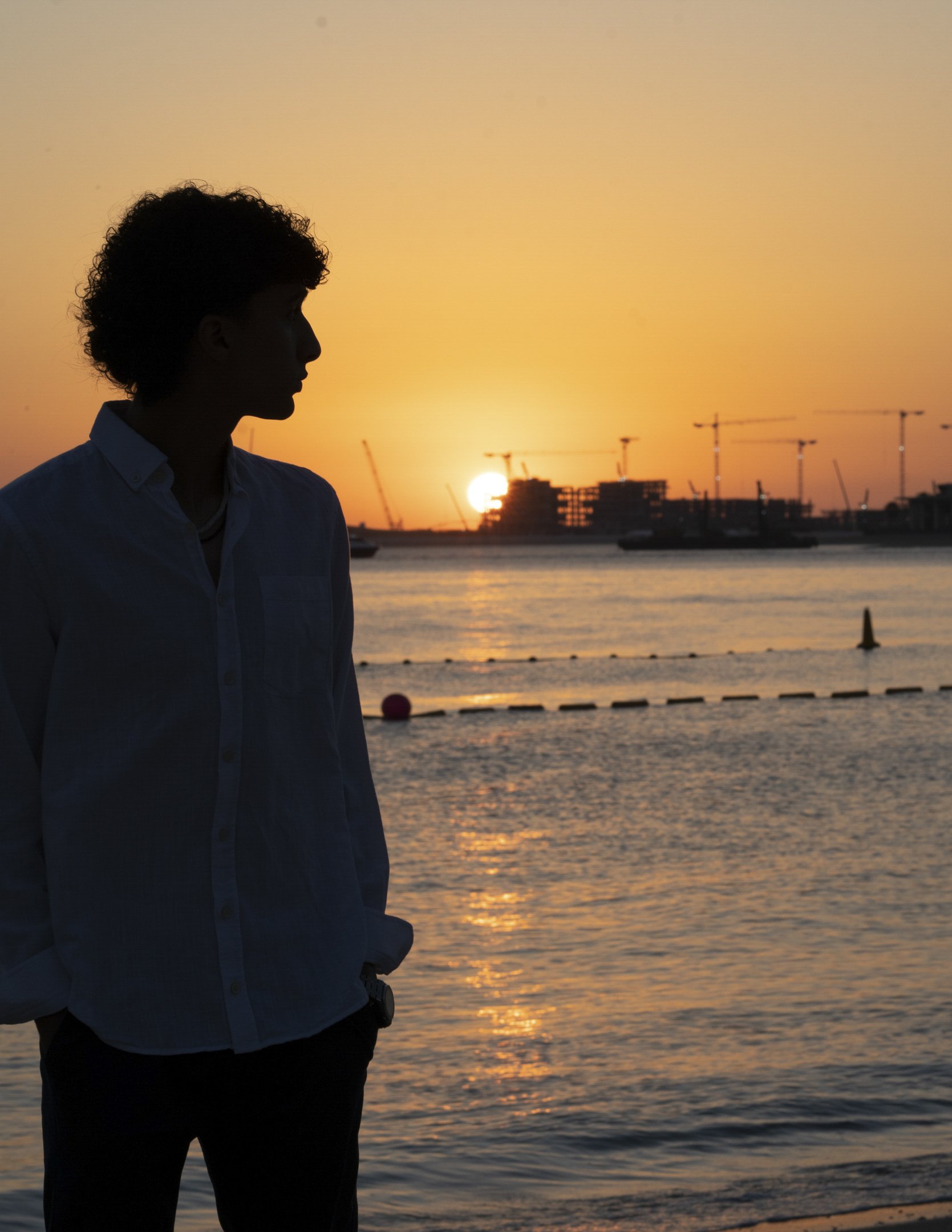 A person in silhouette stands by a body of water during sunset with a construction site and cranes in the background.