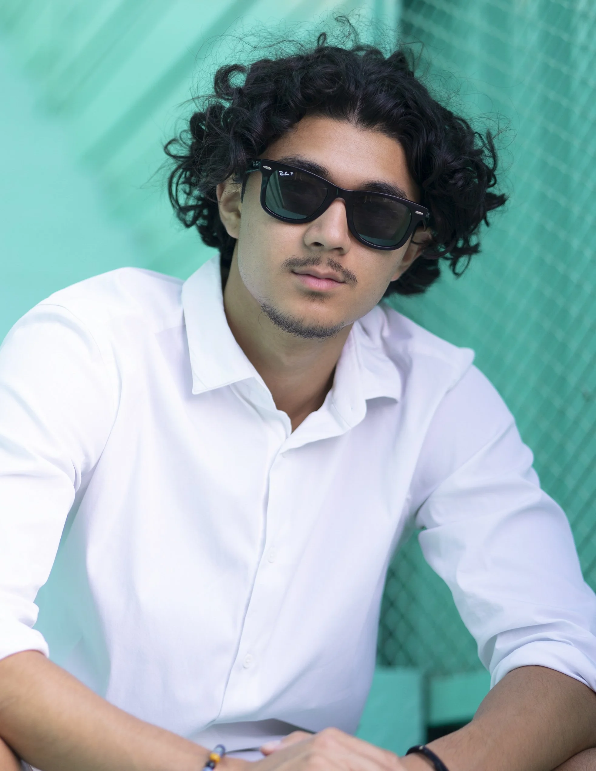 Young man with curly dark hair wearing black sunglasses and a white button-up shirt, sitting against a green and blue background.