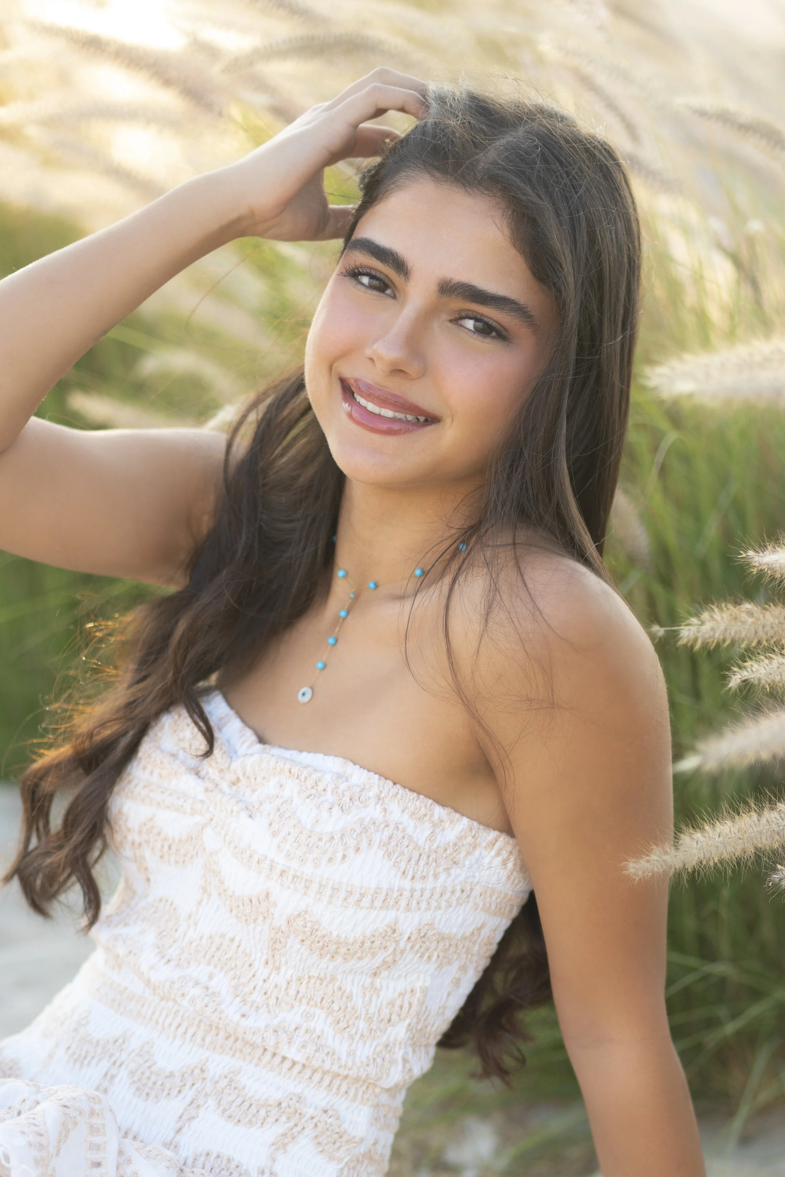A young woman with long brown hair wearing a strapless white dress and a blue beaded necklace, standing outdoors among tall grass and smiling at the camera.