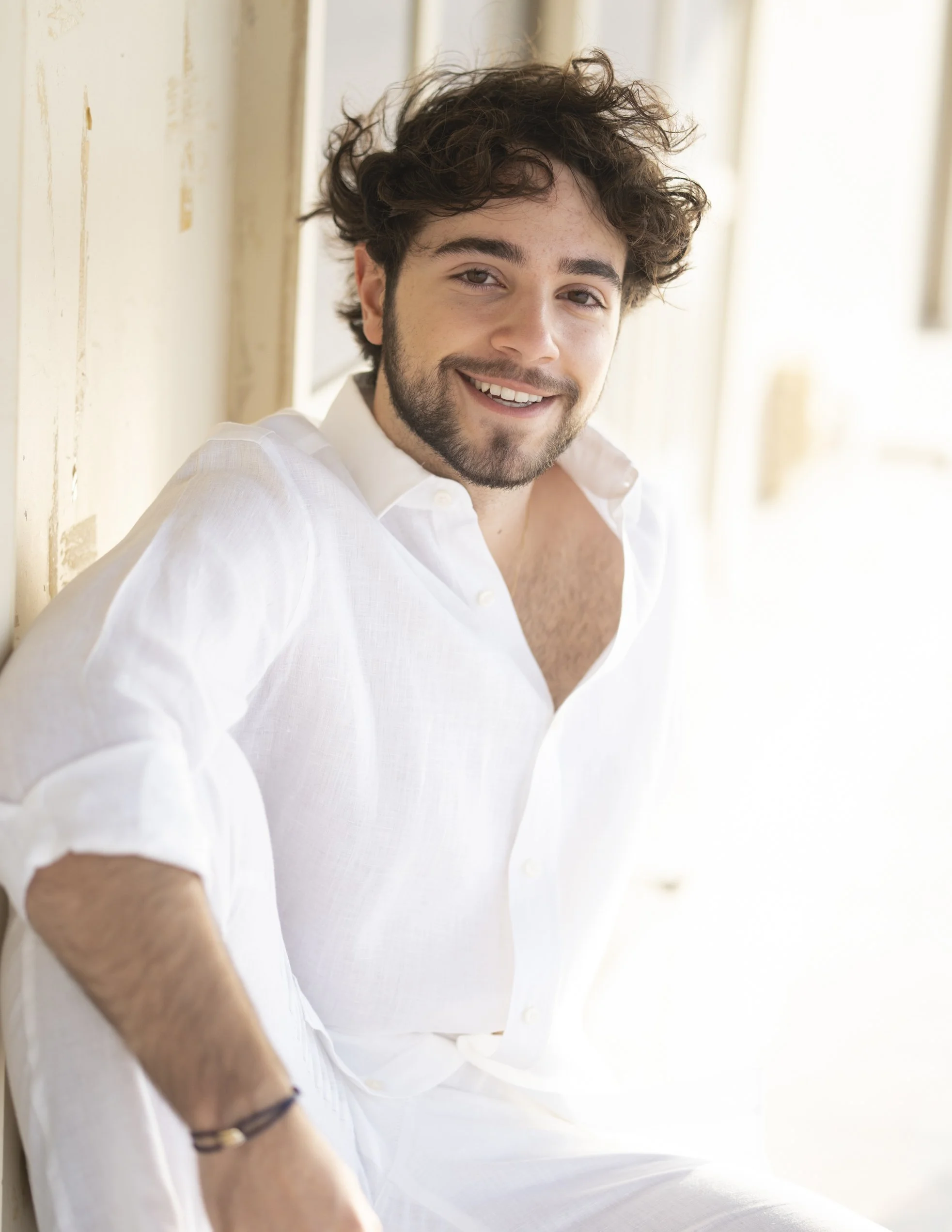 A smiling man with dark curly hair, a beard, and a white shirt sitting against a distressed cream-colored wall.