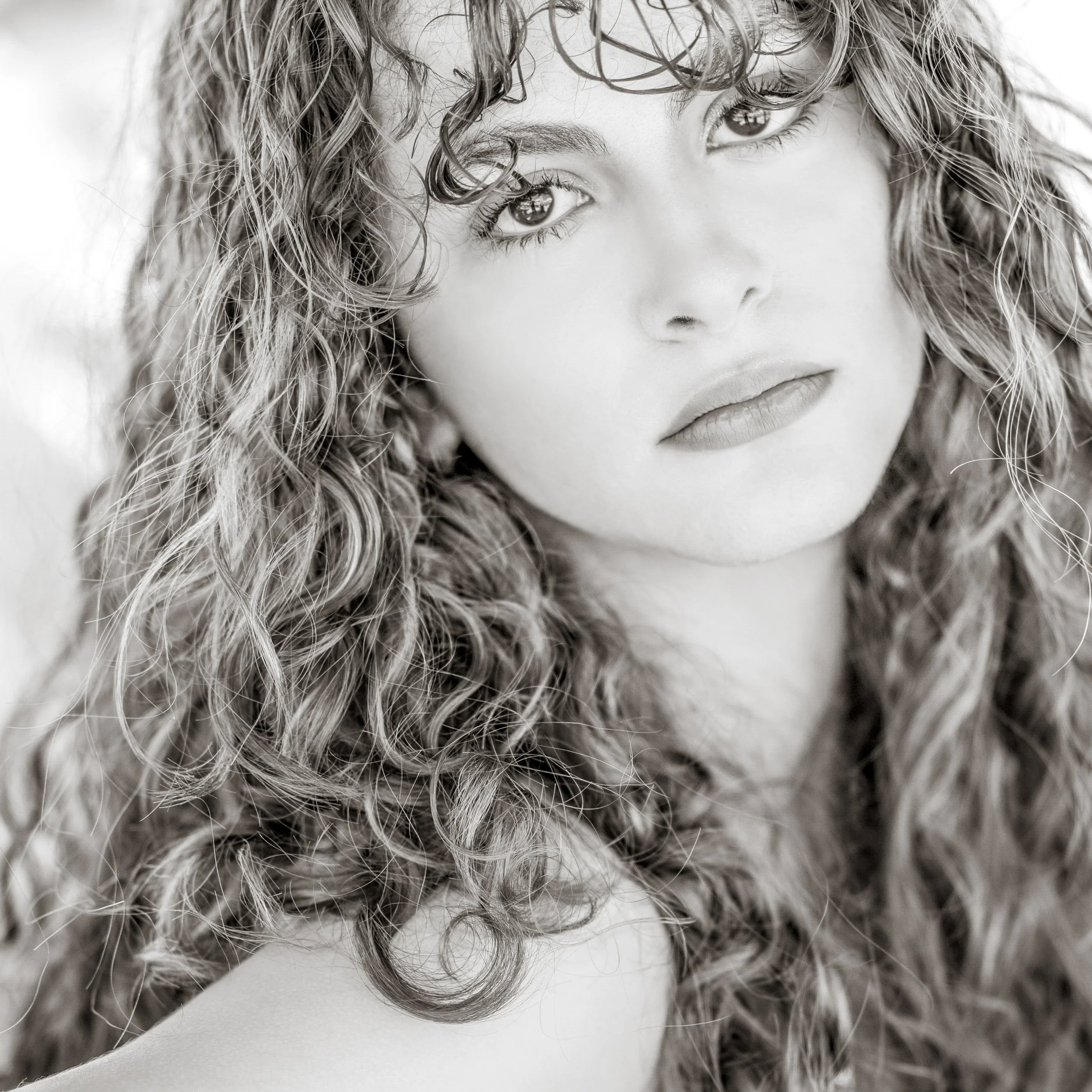 Black and white close-up portrait of a young woman with long curly hair and full lips, looking directly at the camera.