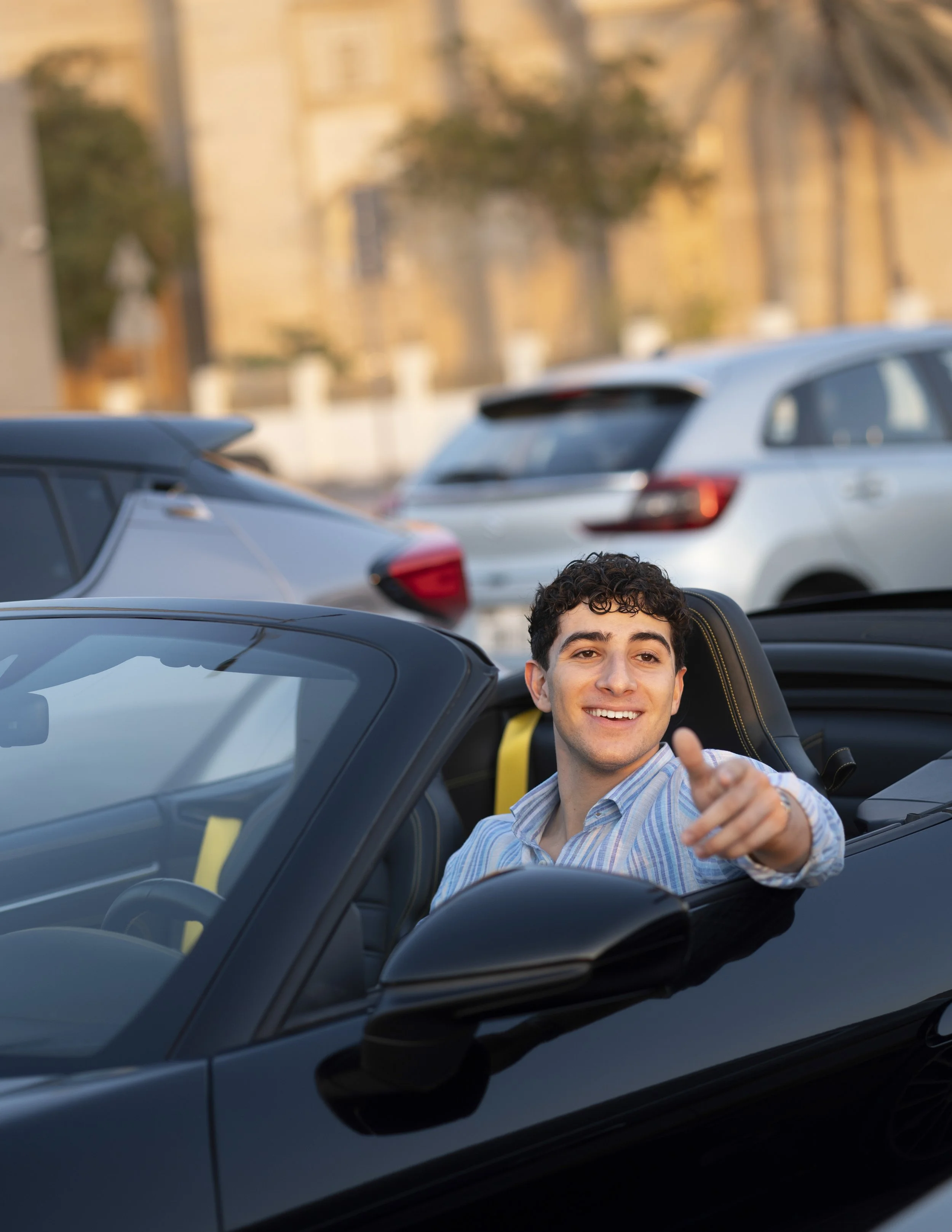 Smiling man in a convertible pointing towards the camera in a parking lot with other cars and buildings in the background.