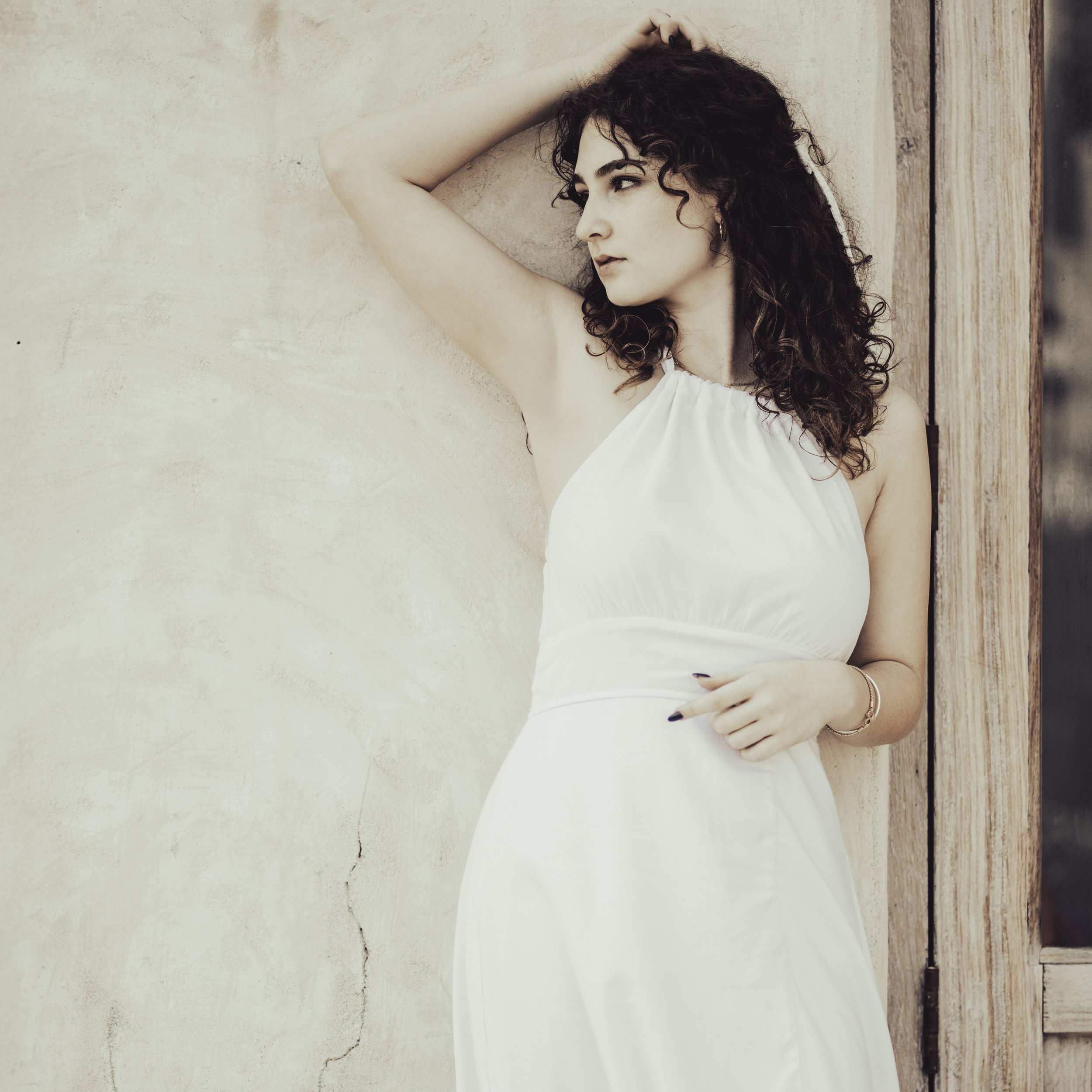 A woman with dark curly hair in a white dress standing against a textured wall with a window frame on the side.