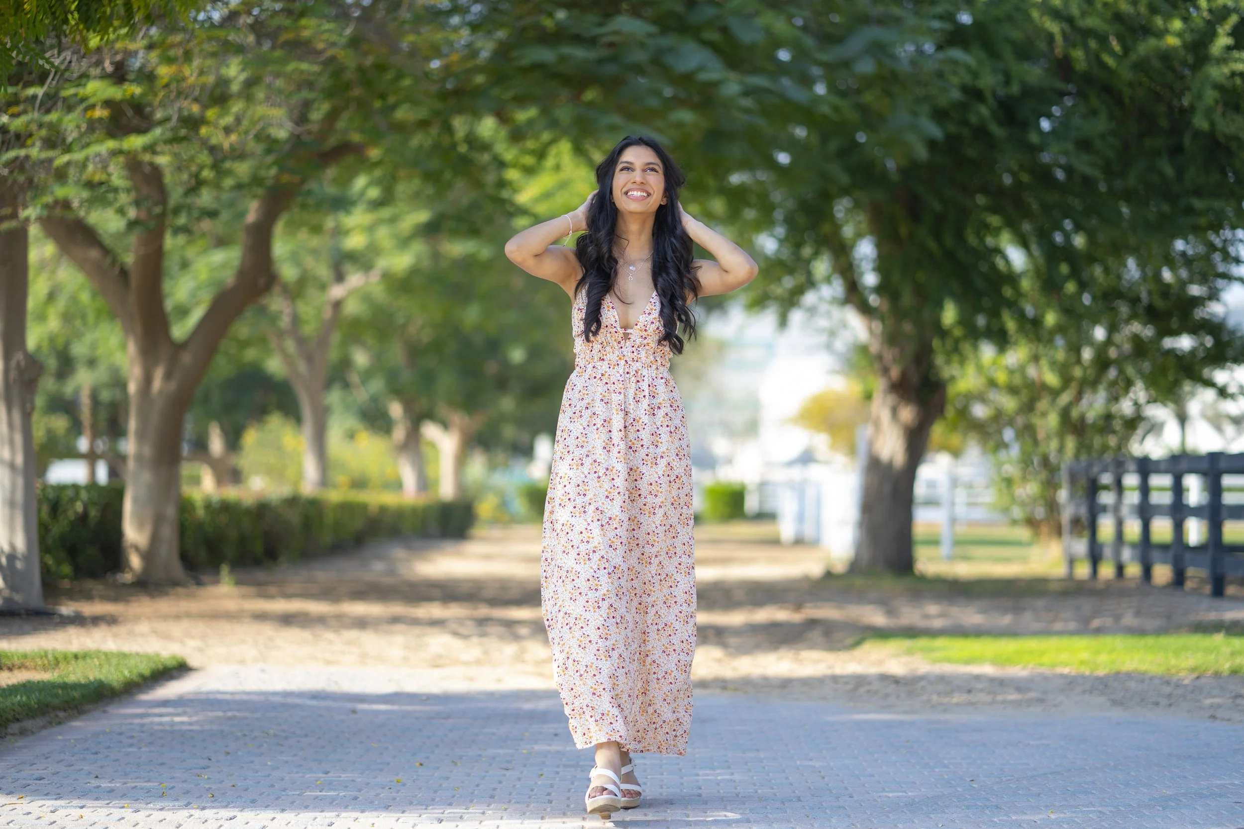 A woman in a floral dress walking on a paved path through a park with trees and greenery, smiling and looking upward.