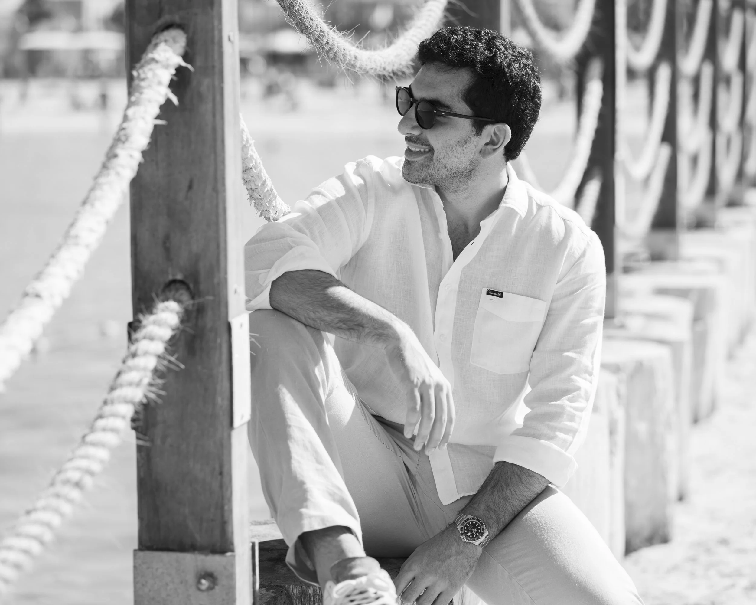 A man wearing sunglasses, a white button-up shirt, and a watch sitting on a wooden bench near a seaside pier, smiling and looking to his right.