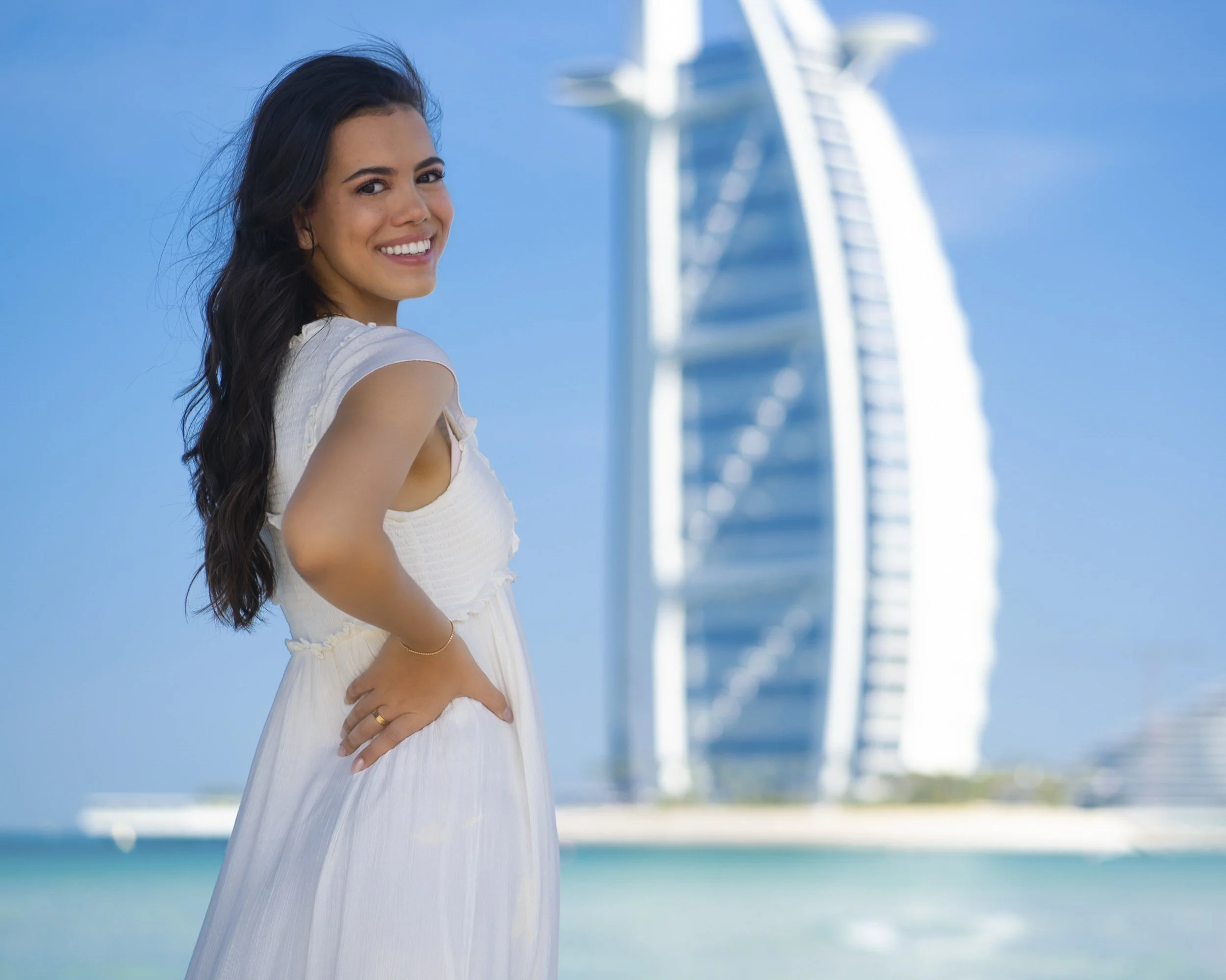 A woman with long dark hair wearing a white dress standing on a beach with the Burj Al Arab hotel in Dubai in the background.