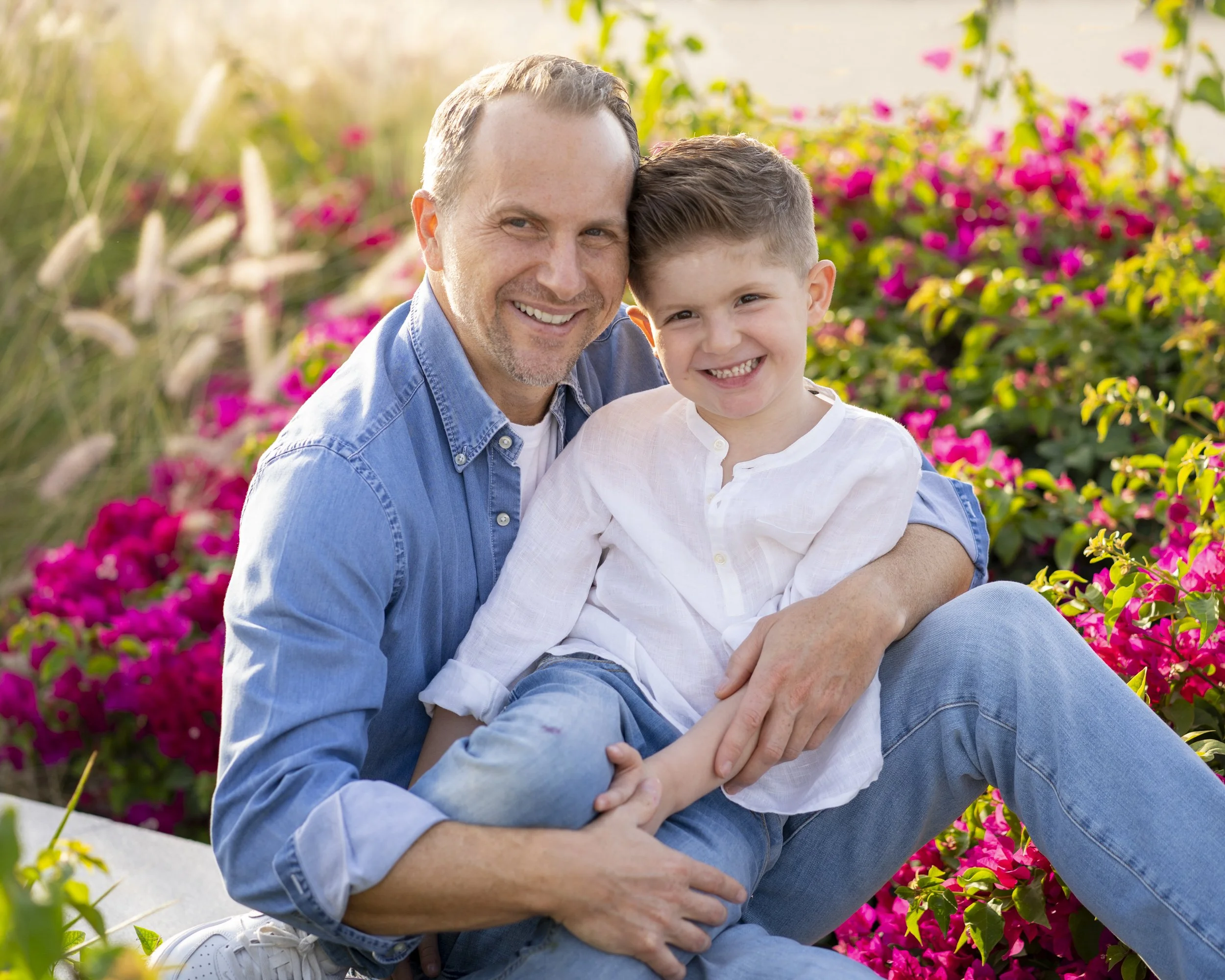 A father and son smiling and sitting together outdoors surrounded by pink and purple flowers.