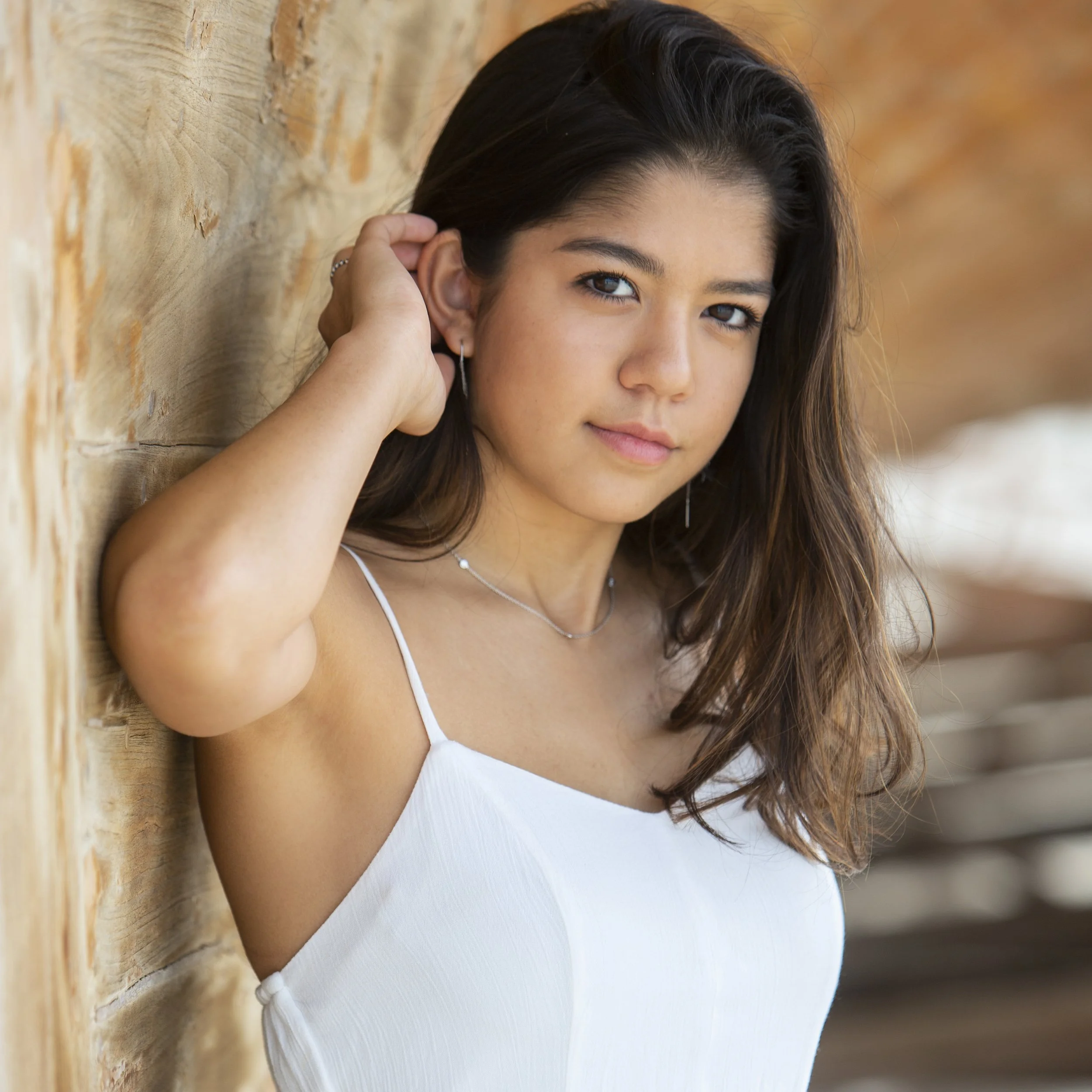A young woman with long brown hair, wearing a white spaghetti strap top and jewelry, leaning against a wooden wall outdoors.