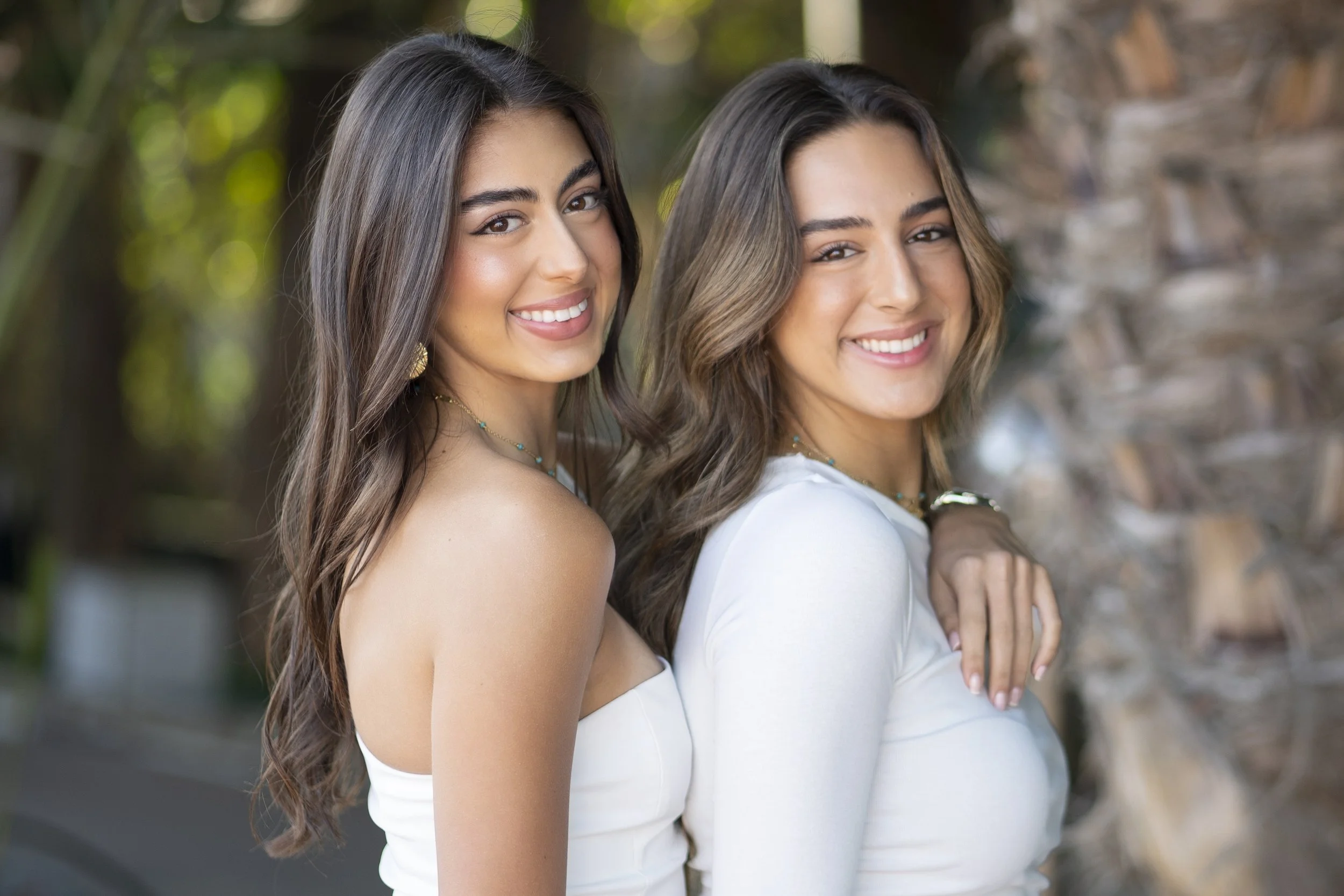 Two young women with long brown hair and light skin smiling outdoors in front of a blurred background with greenery and a tree trunk. They are close to each other, facing the camera, with one resting her hand on the other's shoulder, both wearing whi